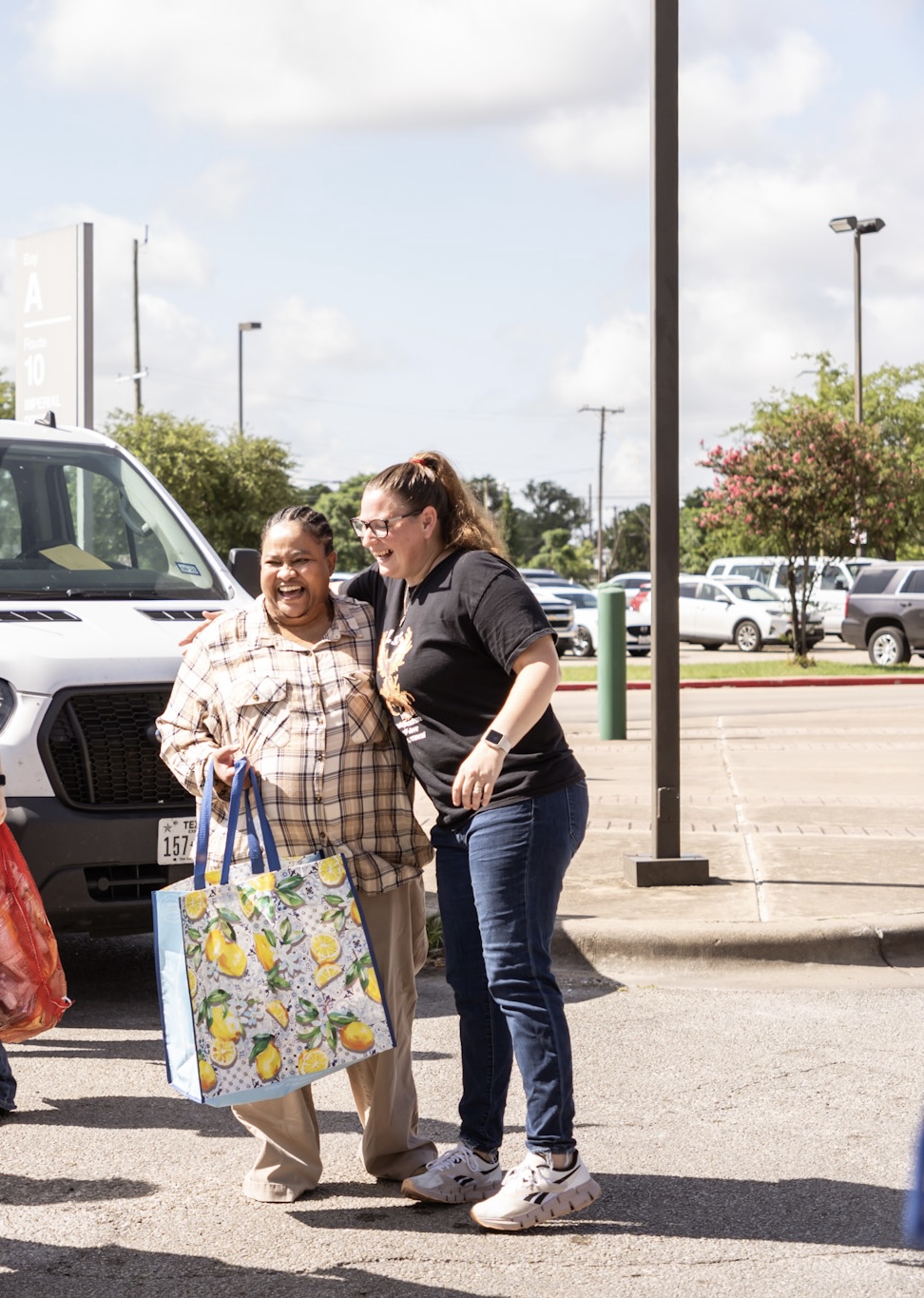 Two women smiling and talking in a parking lot, one holding a colorful bag, with cars and trees in the background.