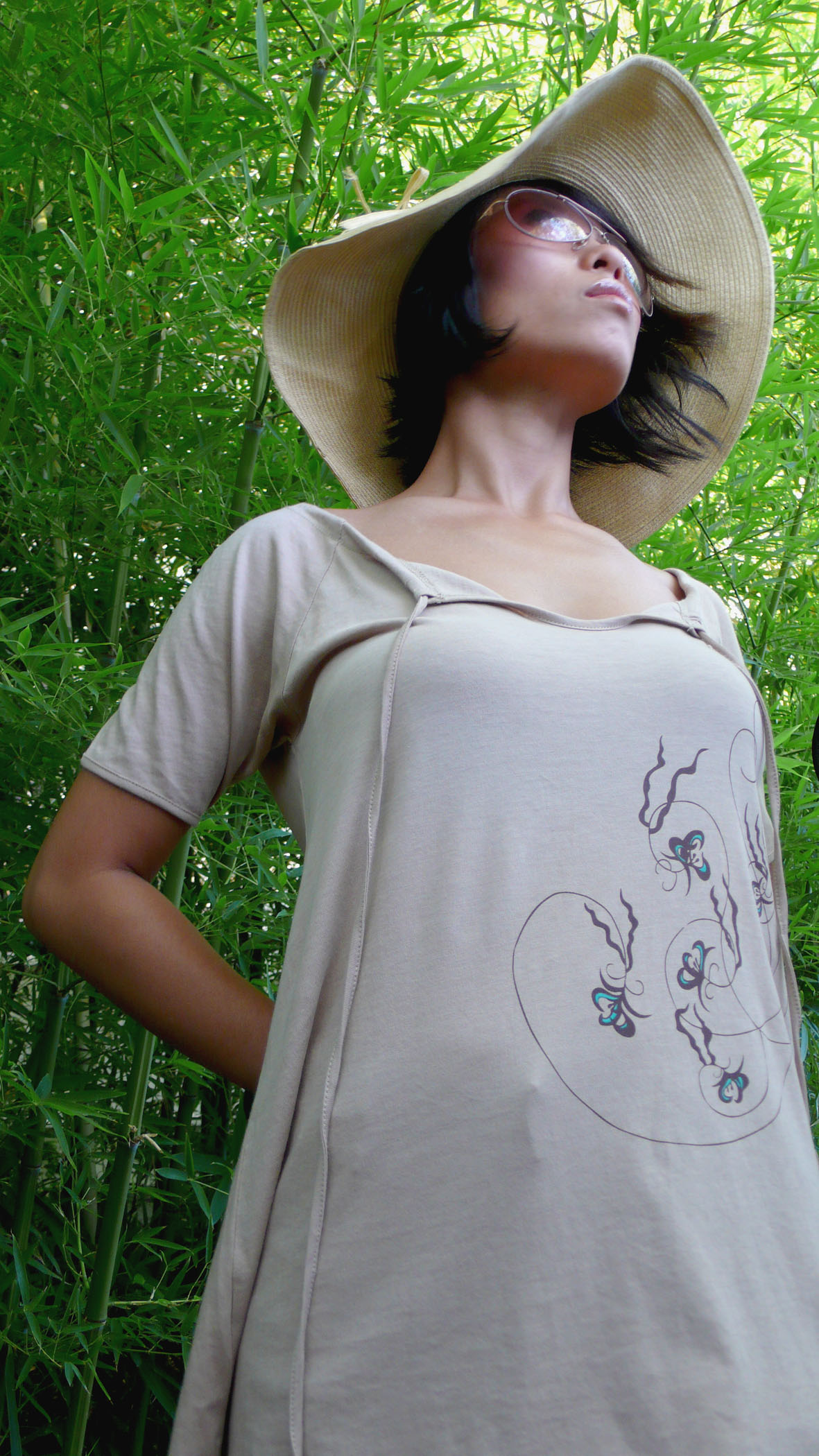Woman wearing large hat and sunglasses, standing outdoors among green plants, looking upward, with a drawing of a heart and faces on her shirt.