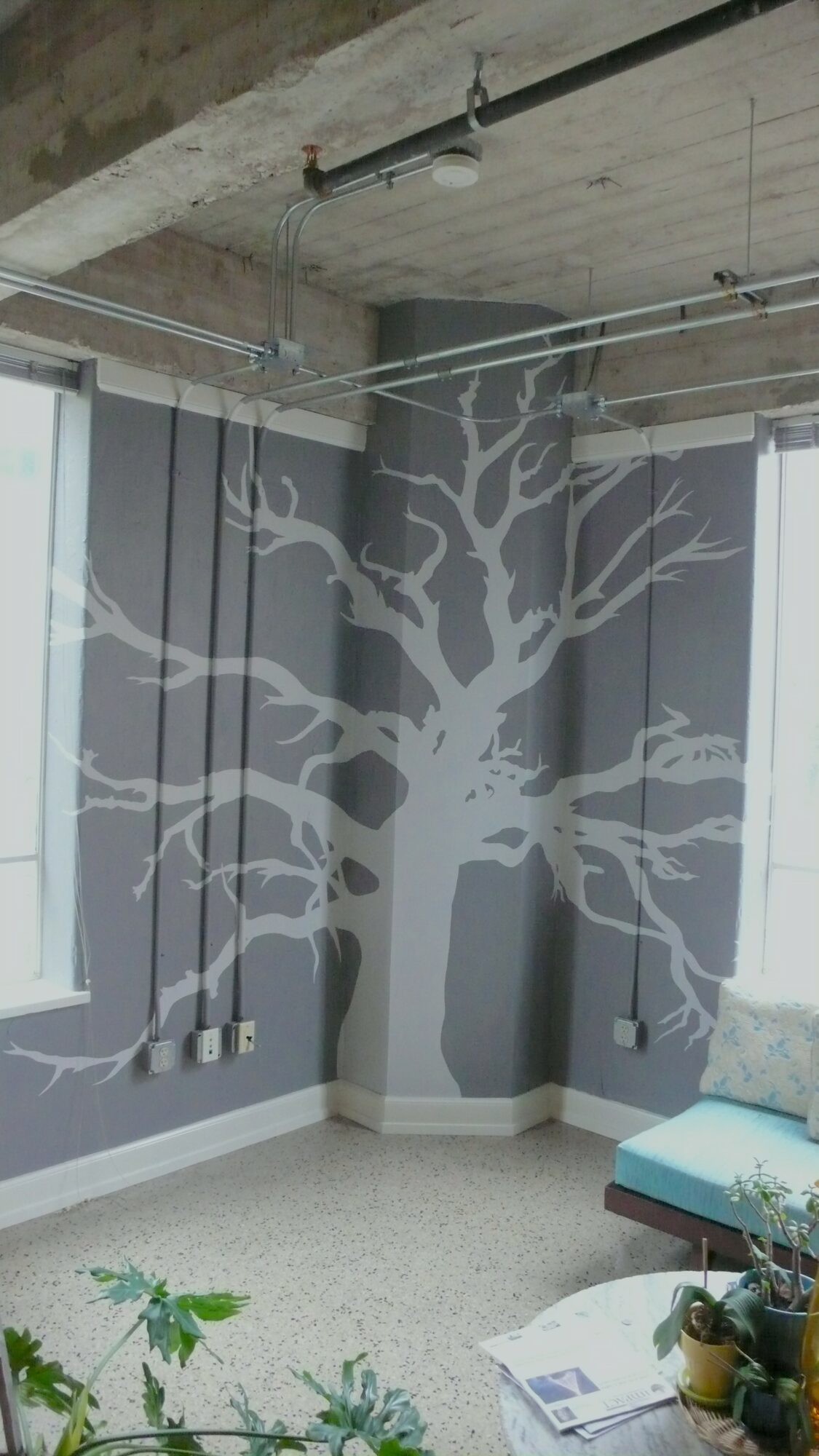 Interior corner with a large tree mural on the wall, windows with blinds, and a blue sofa with a pillow.