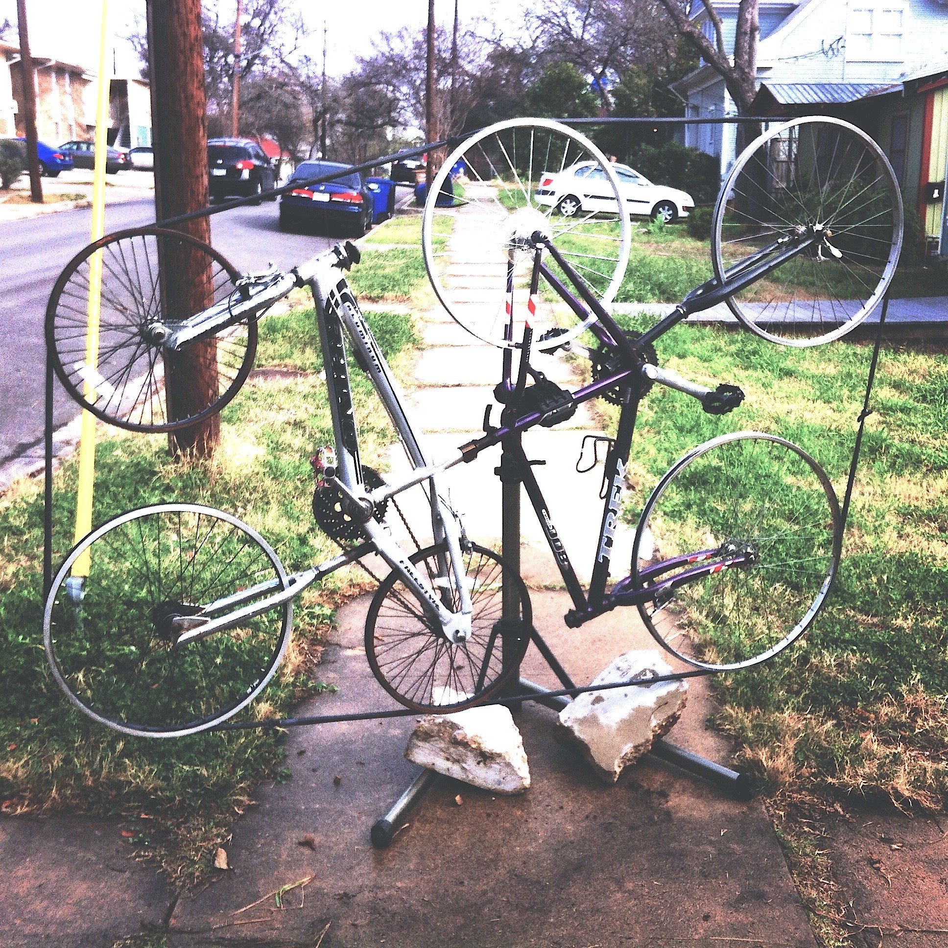 Bicycle sculpture made of metal and rocks, mounted outdoors on a sidewalk with grass and trees in background.