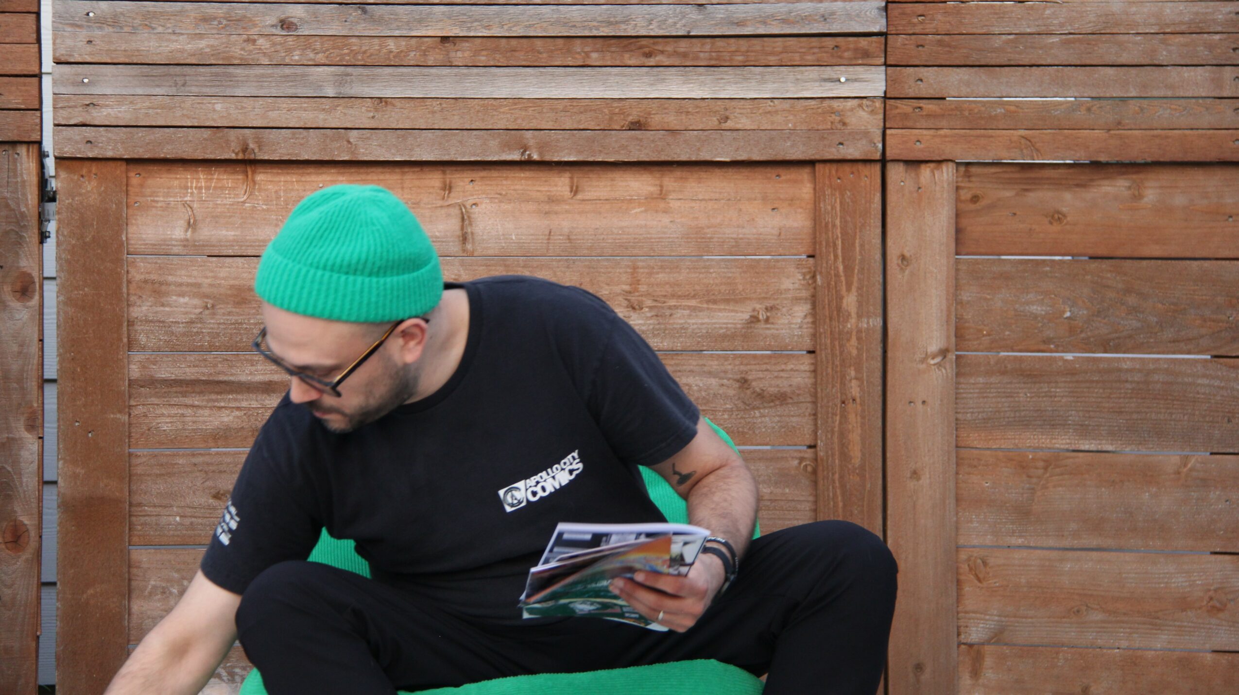 Person wearing a turquoise beanie and black shirt sitting outdoors in front of a wooden fence, holding a magazine.