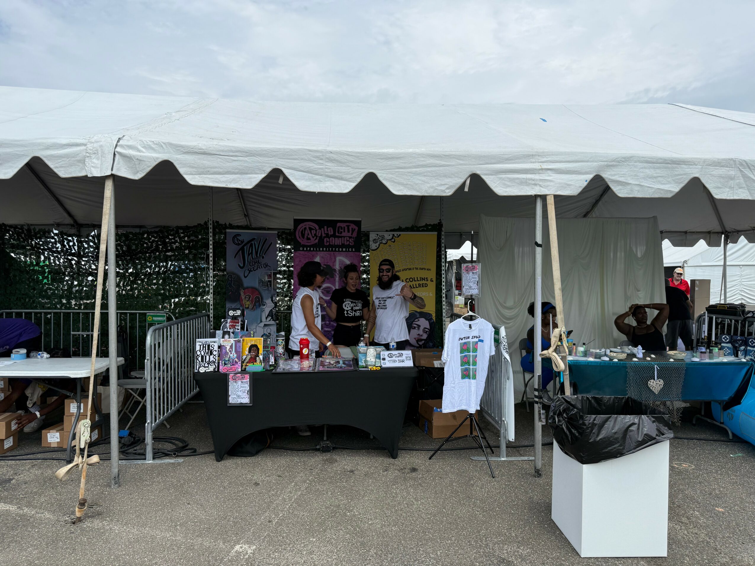 People are gathered under a tent at an outdoor event, with tables displaying items and promotional posters behind them.