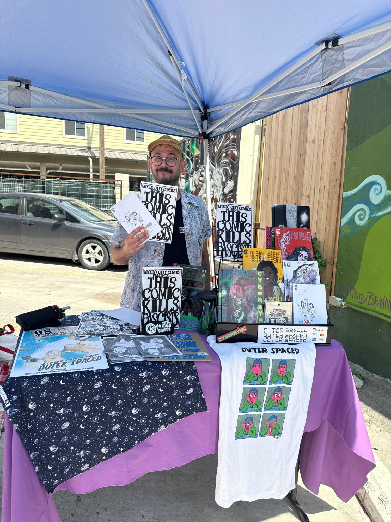 Person holding a book at an outdoor market stall with various items, including books and fabric, under a blue canopy.