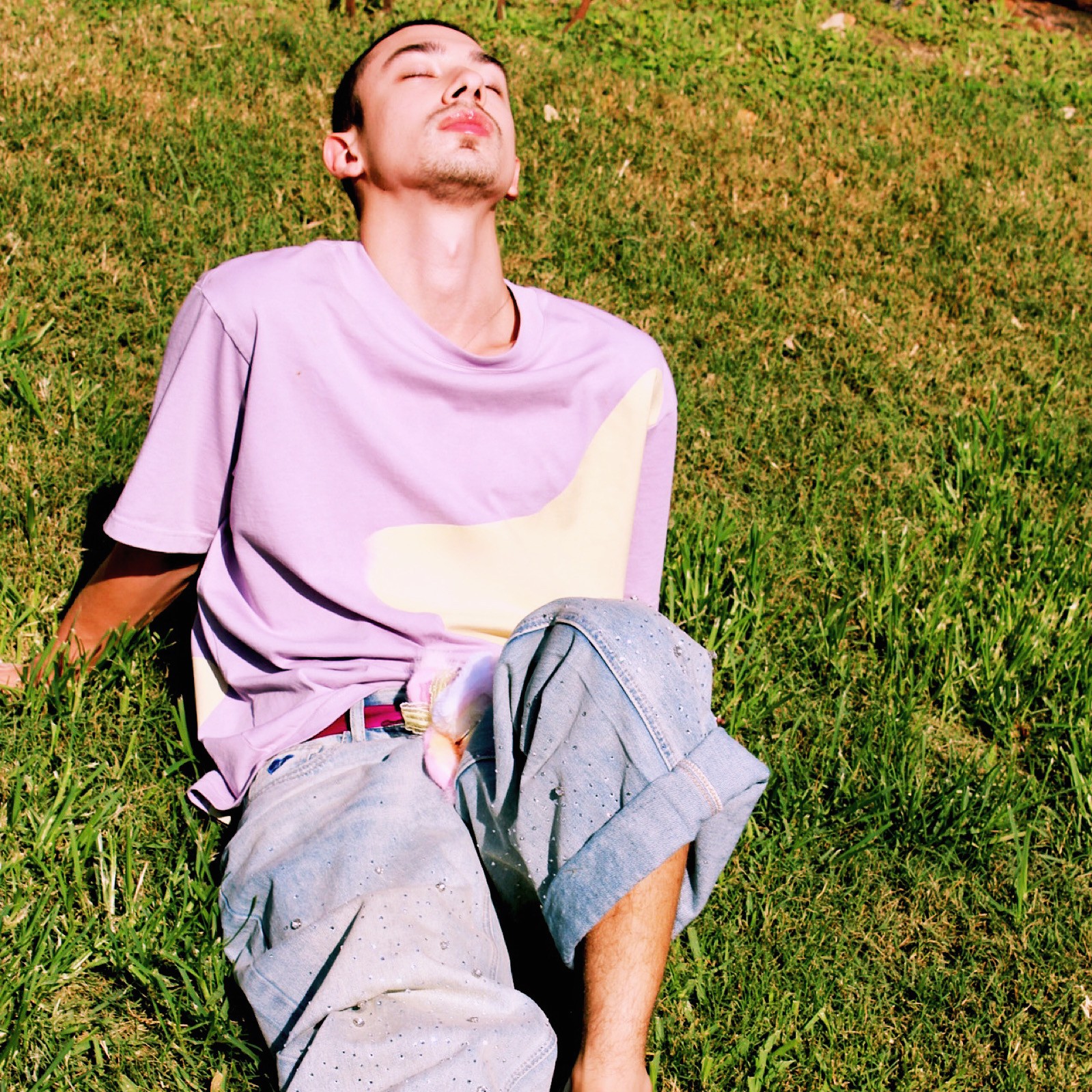 Person sitting on grass with eyes closed, wearing a light purple shirt and gray pants, relaxing outdoors.