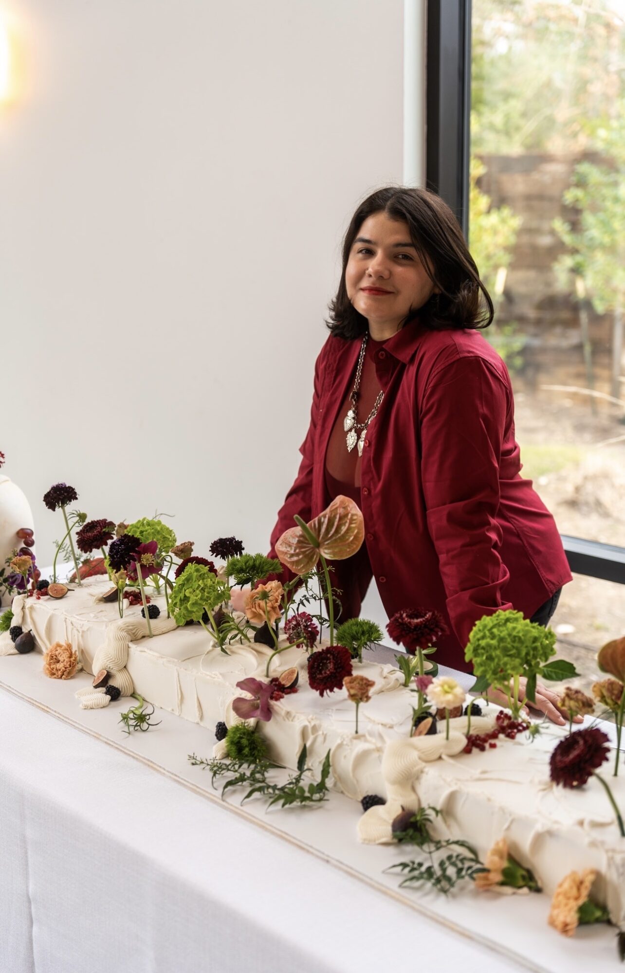 Woman with dark hair wearing a red jacket stands behind a table with decorated cakes, near a window with trees outside.