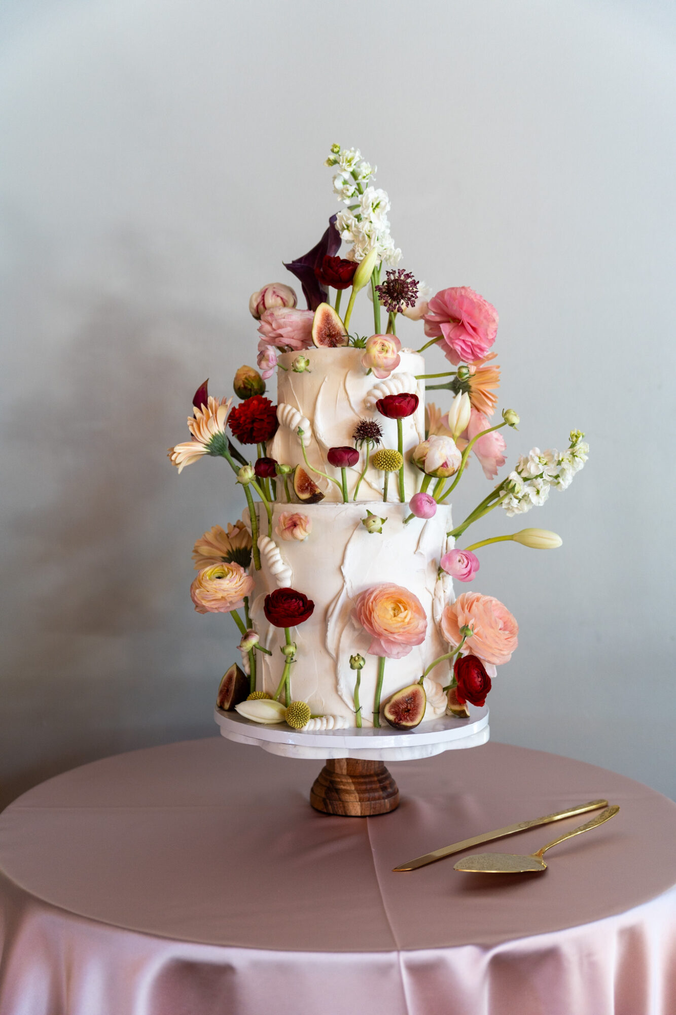 Three-tiered white cake decorated with pink, red, and white flowers, topped with a small bouquet, on a cake stand.