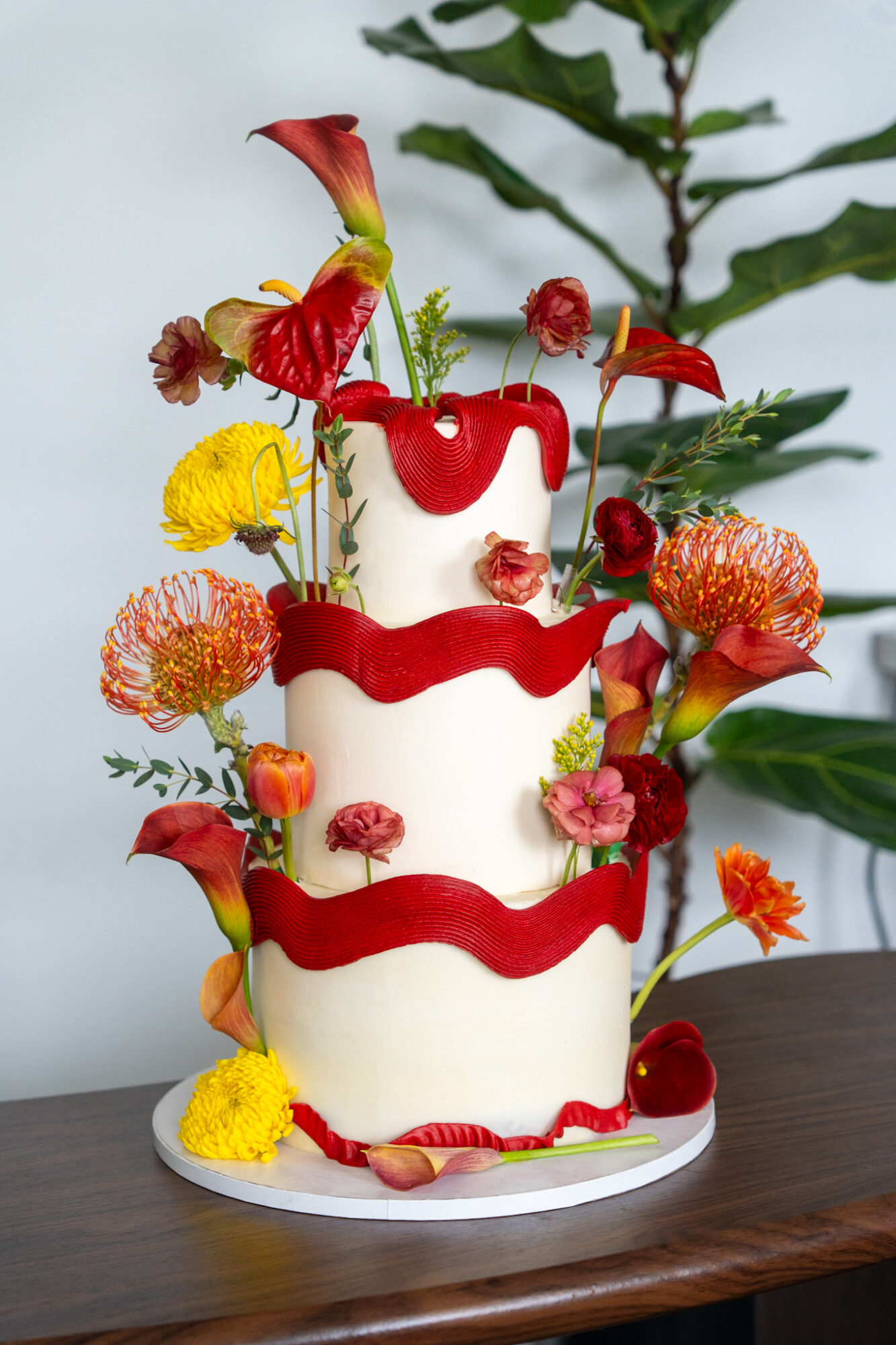 Three-tiered cake decorated with red, yellow, and pink flowers, and green foliage, on a wooden surface.