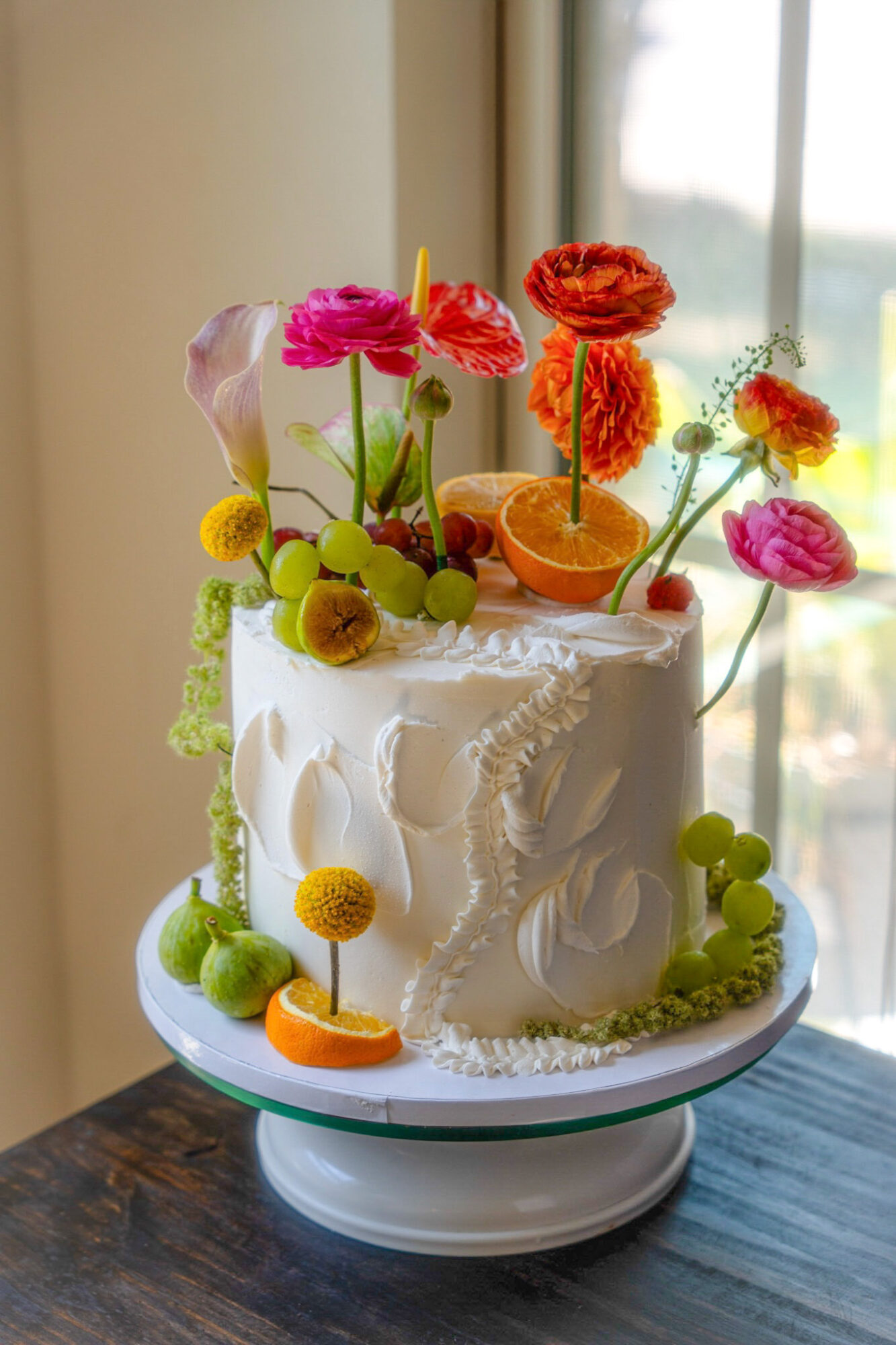 Decorated cake with colorful flowers and fruit slices on top, placed on a white cake stand near a window.