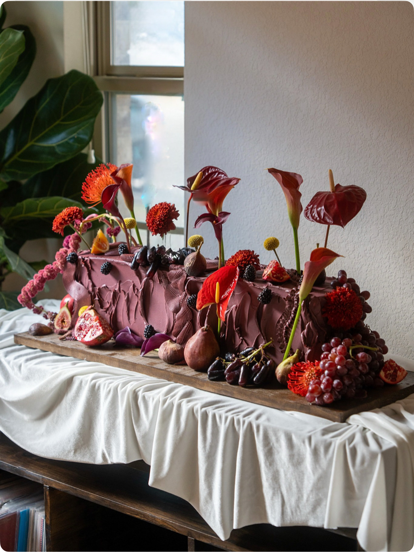 Decorative arrangement of red and pink flowers and berries on a table with a white tablecloth, near a window.
