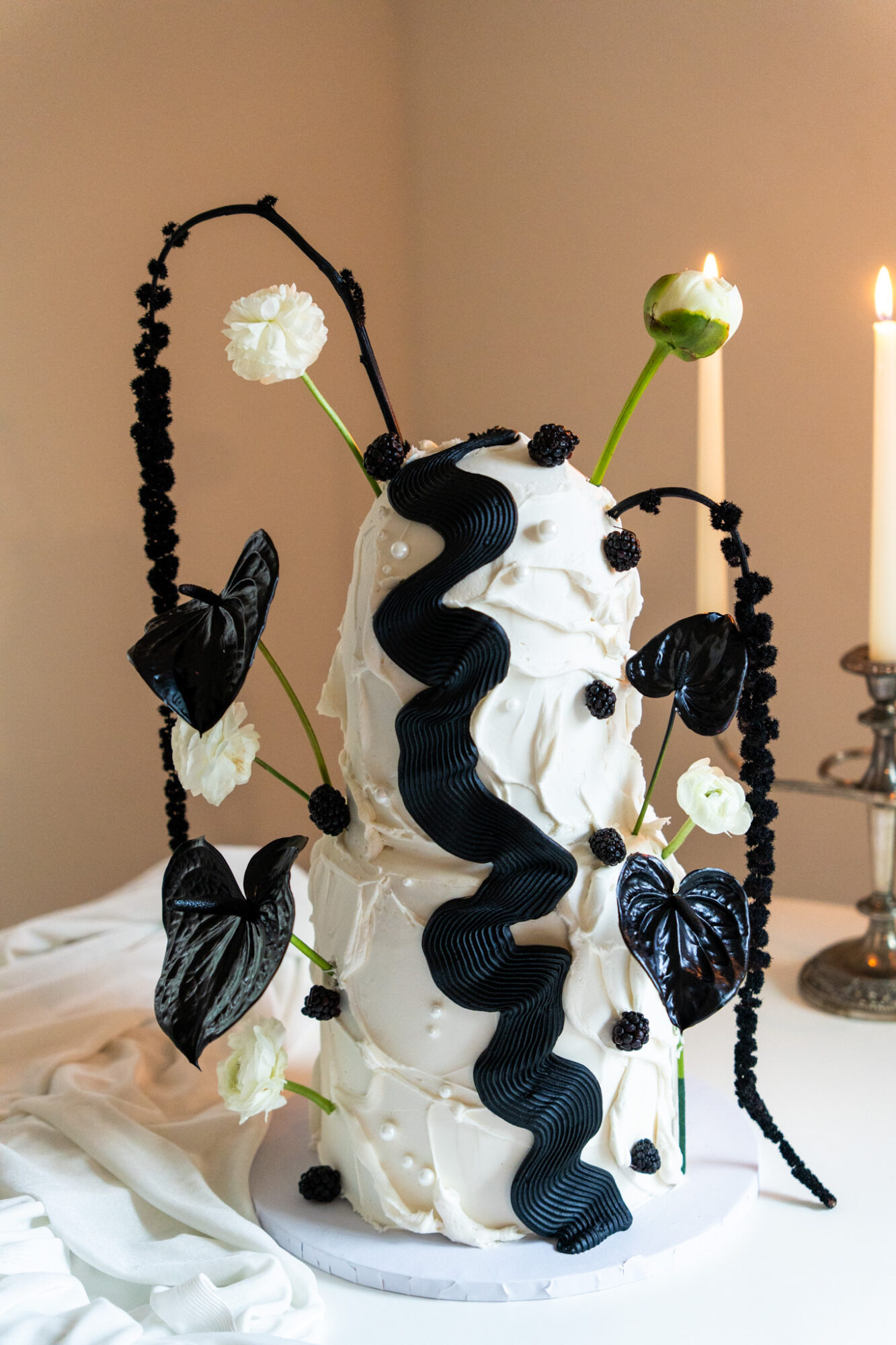 Decorative cake with black and white elements, flowers, and black butterflies, on a table with a candle in the background.