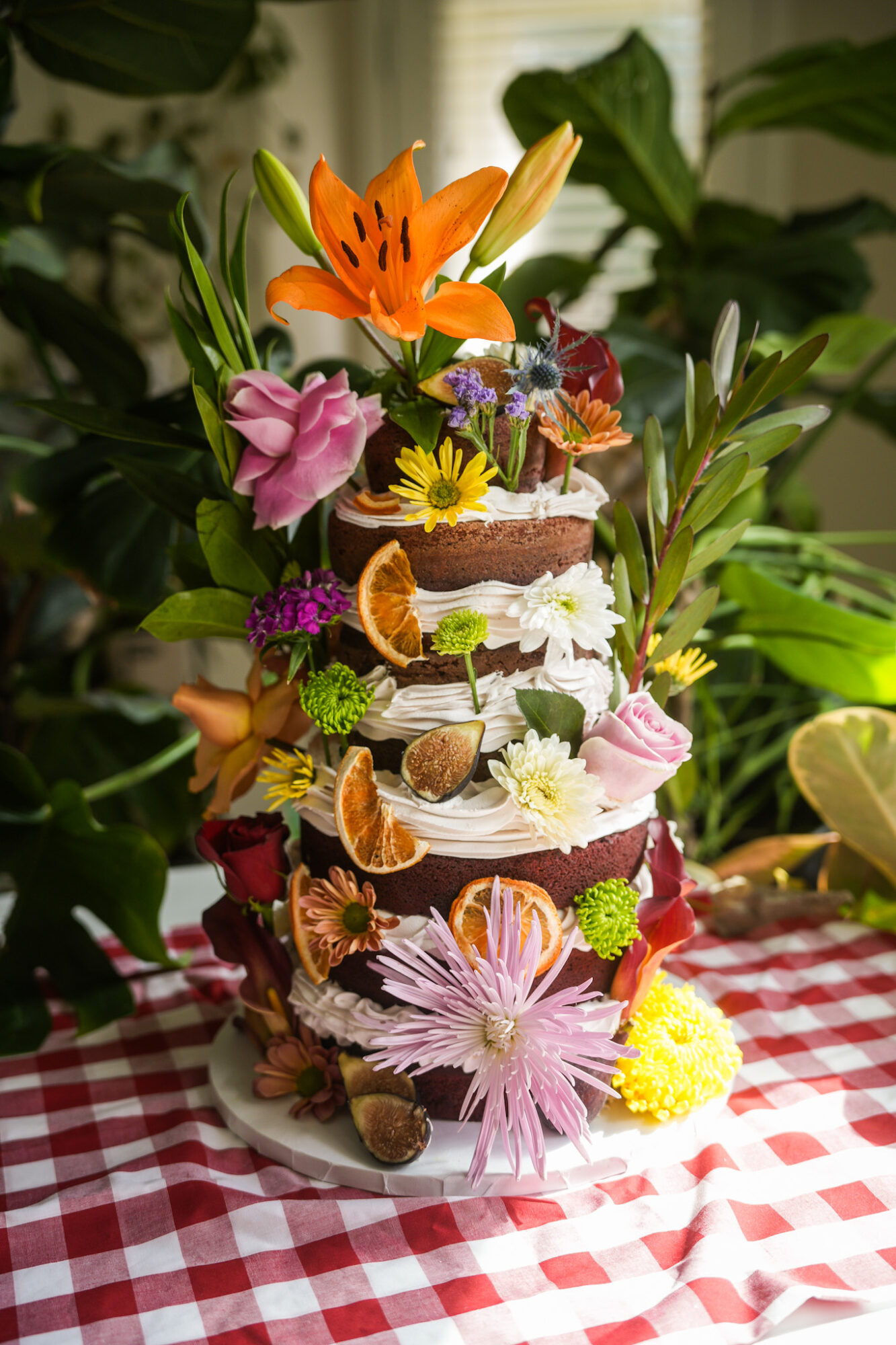 Multi-tiered cake decorated with flowers, fruit slices, and greenery, placed on a red and white checkered tablecloth, with plants in background.