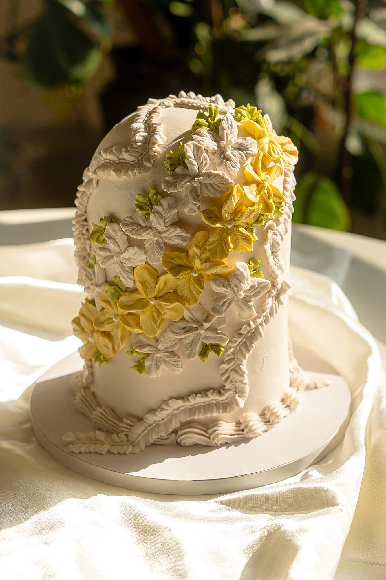 Decorative cake shaped like a boot with white and yellow flowers, on a white cloth, with green plants in background.