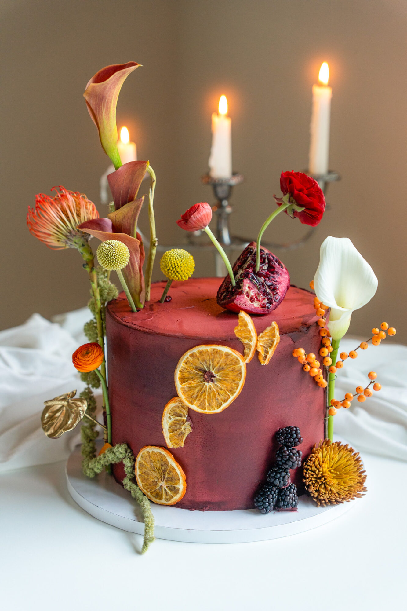 A decorated cake with flowers, dried citrus slices, berries, and three lit candles on top.