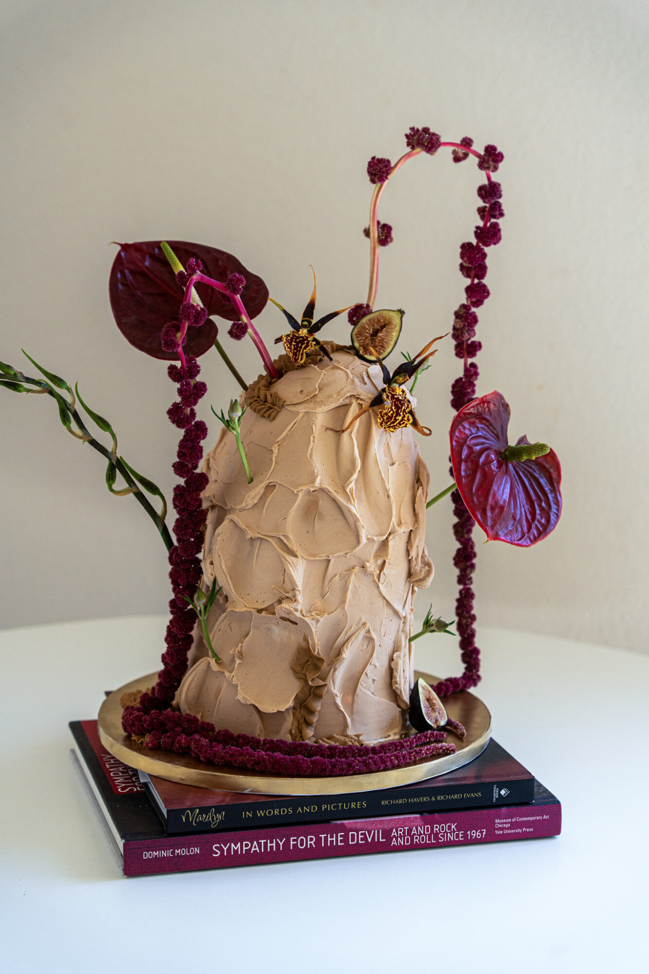Decorative sculpture with textured beige surface, surrounded by purple and green plants, placed on two stacked books, against a plain background.