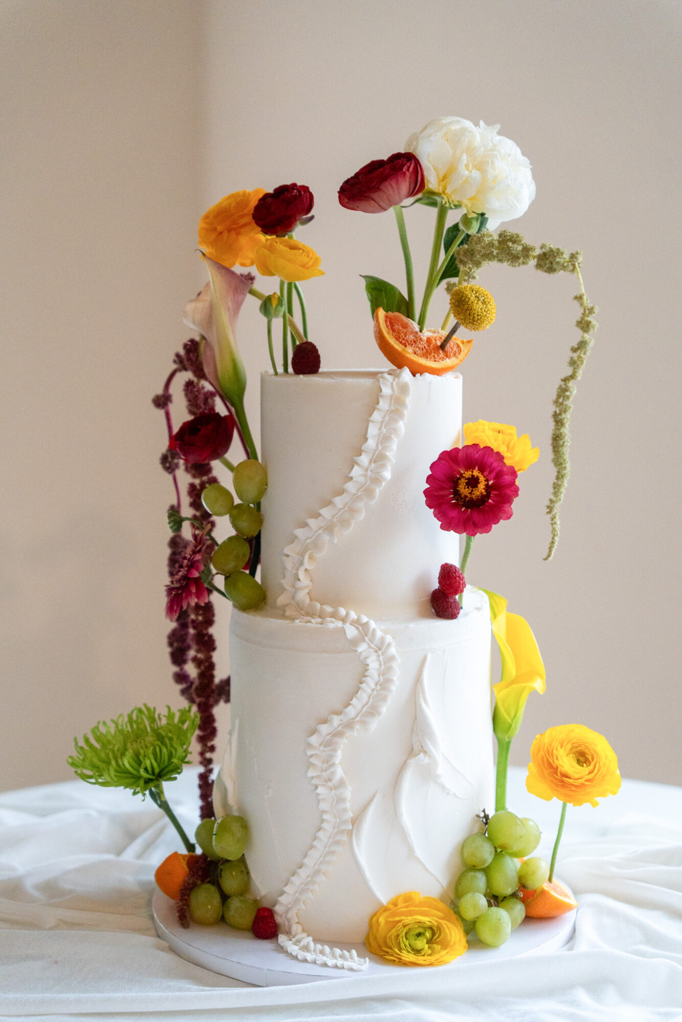 Two-tiered white cake decorated with colorful flowers and fruit, with a neutral background.