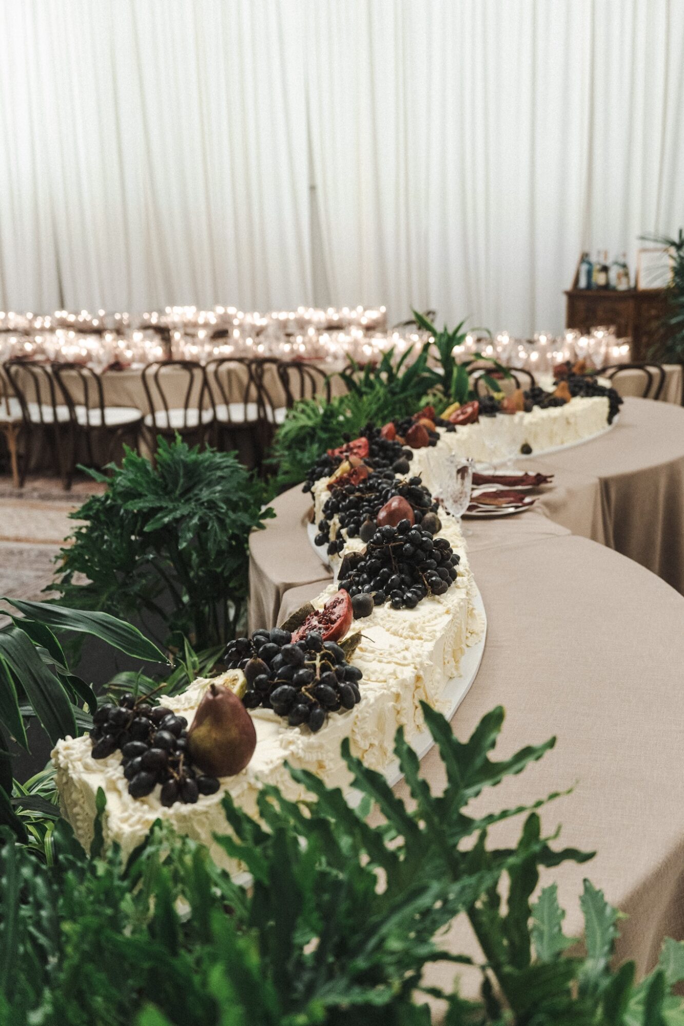 Decorated cake with berries and greenery on a table, with chairs and tables in the background, in a decorated event space.