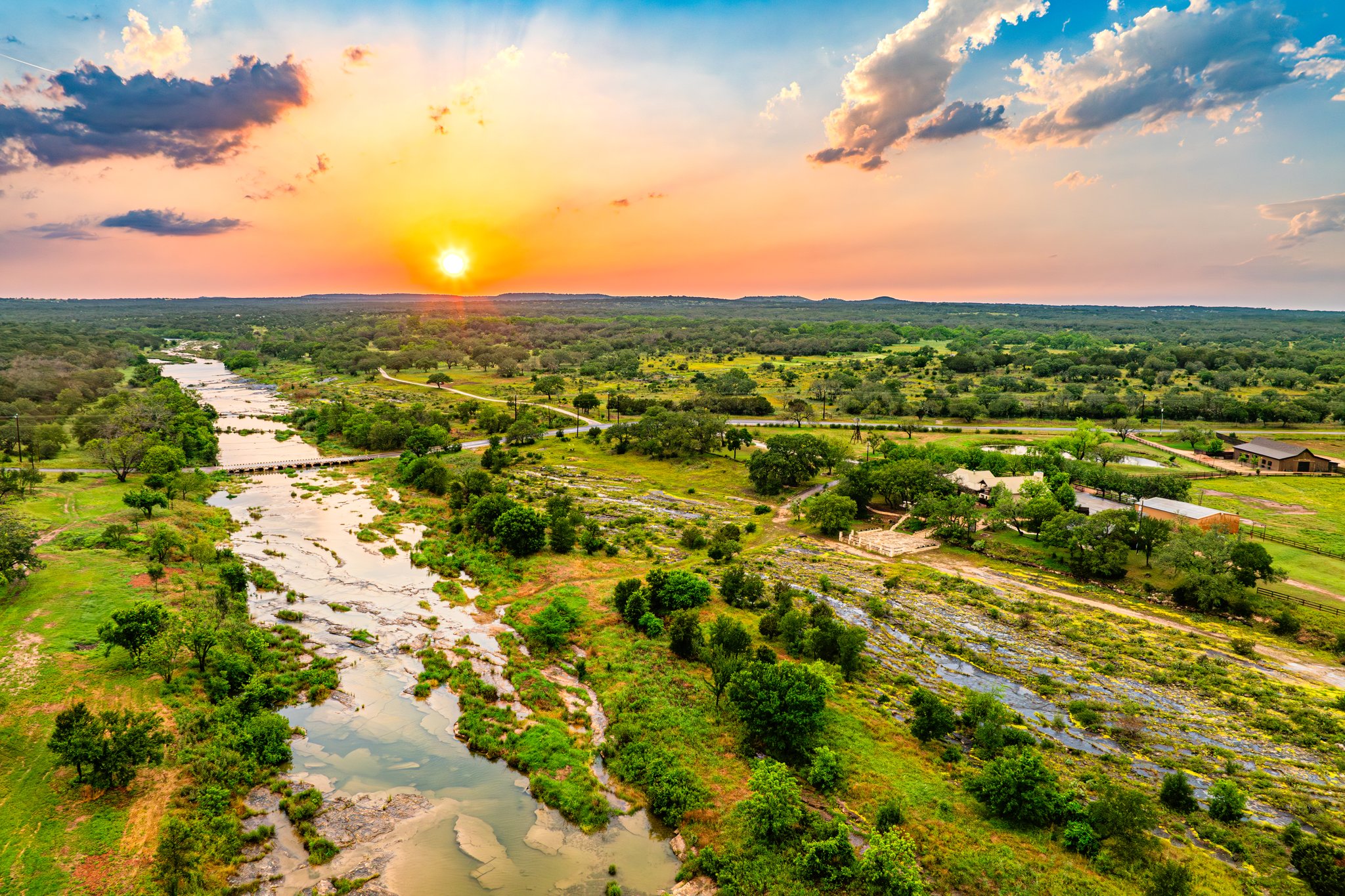 A landscape with a river flowing through green fields and trees, sunset sky with clouds, and distant horizon.
