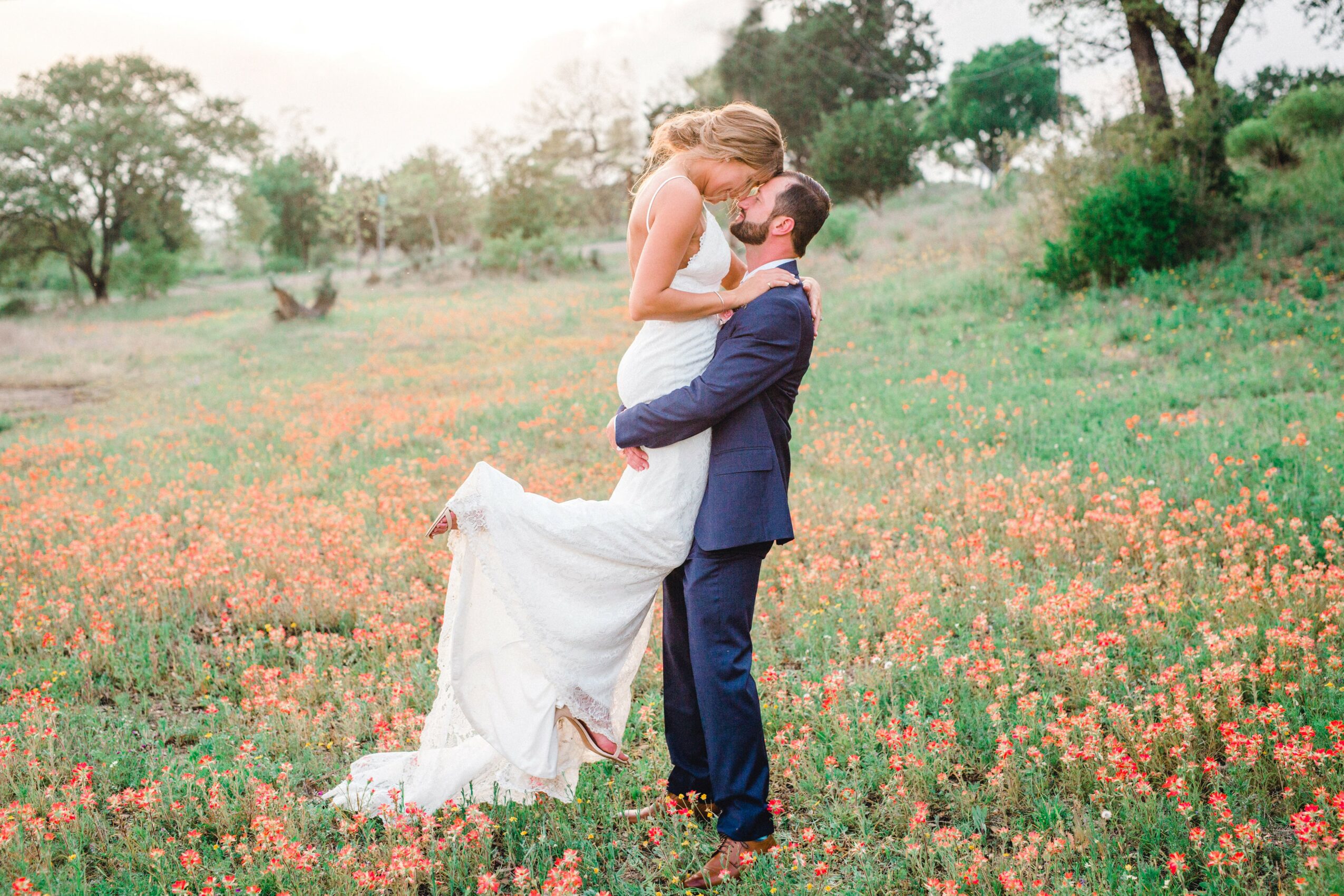 A couple in wedding attire embracing in a field of pink flowers with trees in the background.