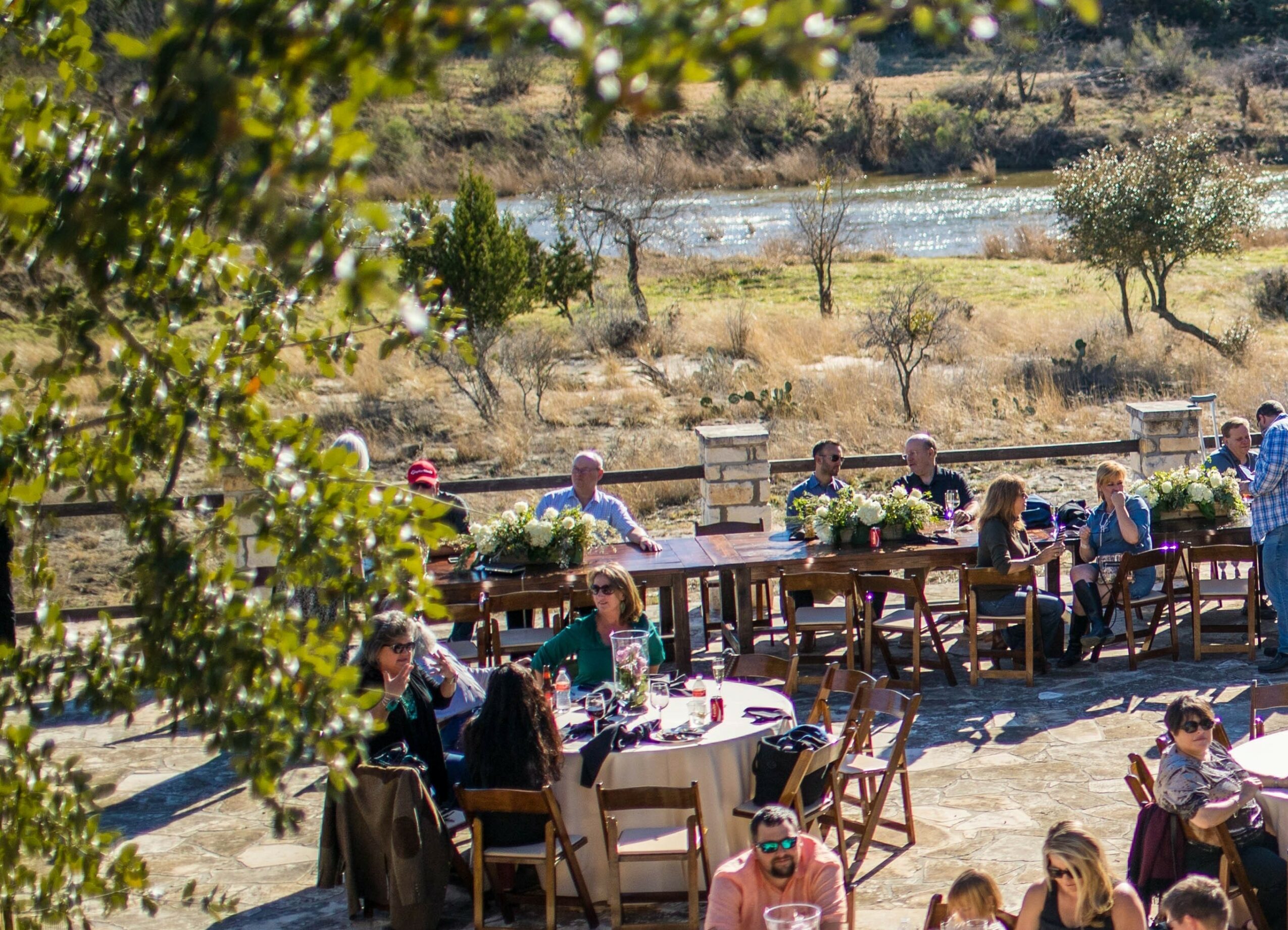 People dining outdoors at tables with a scenic landscape of trees, water, and hills in the background.