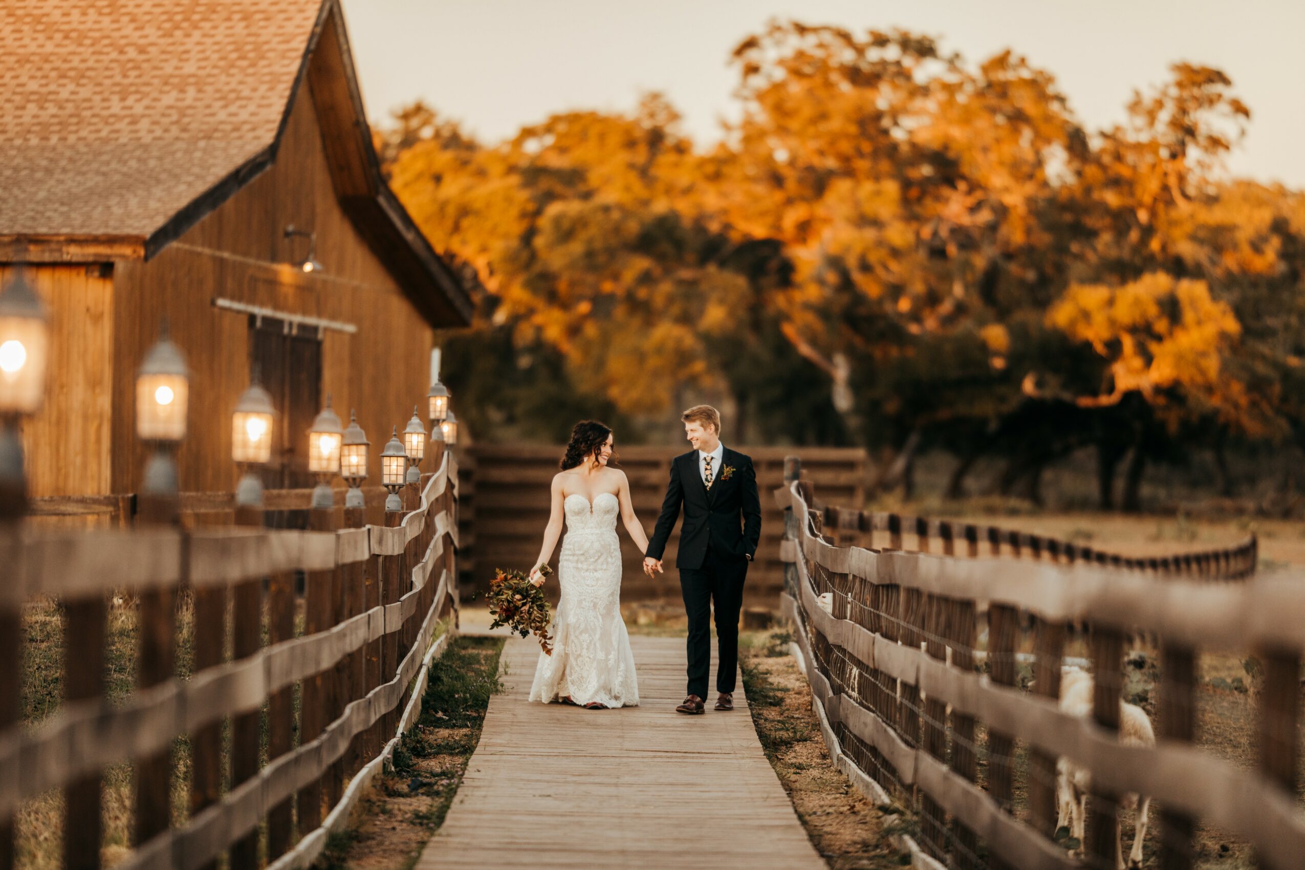 A bride and groom walk hand in hand on a wooden bridge outdoors during sunset, with trees in the background.