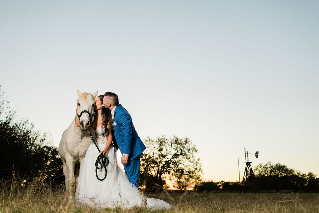 Person in wedding dress and person in blue suit kissing next to a white horse outdoors at sunset.