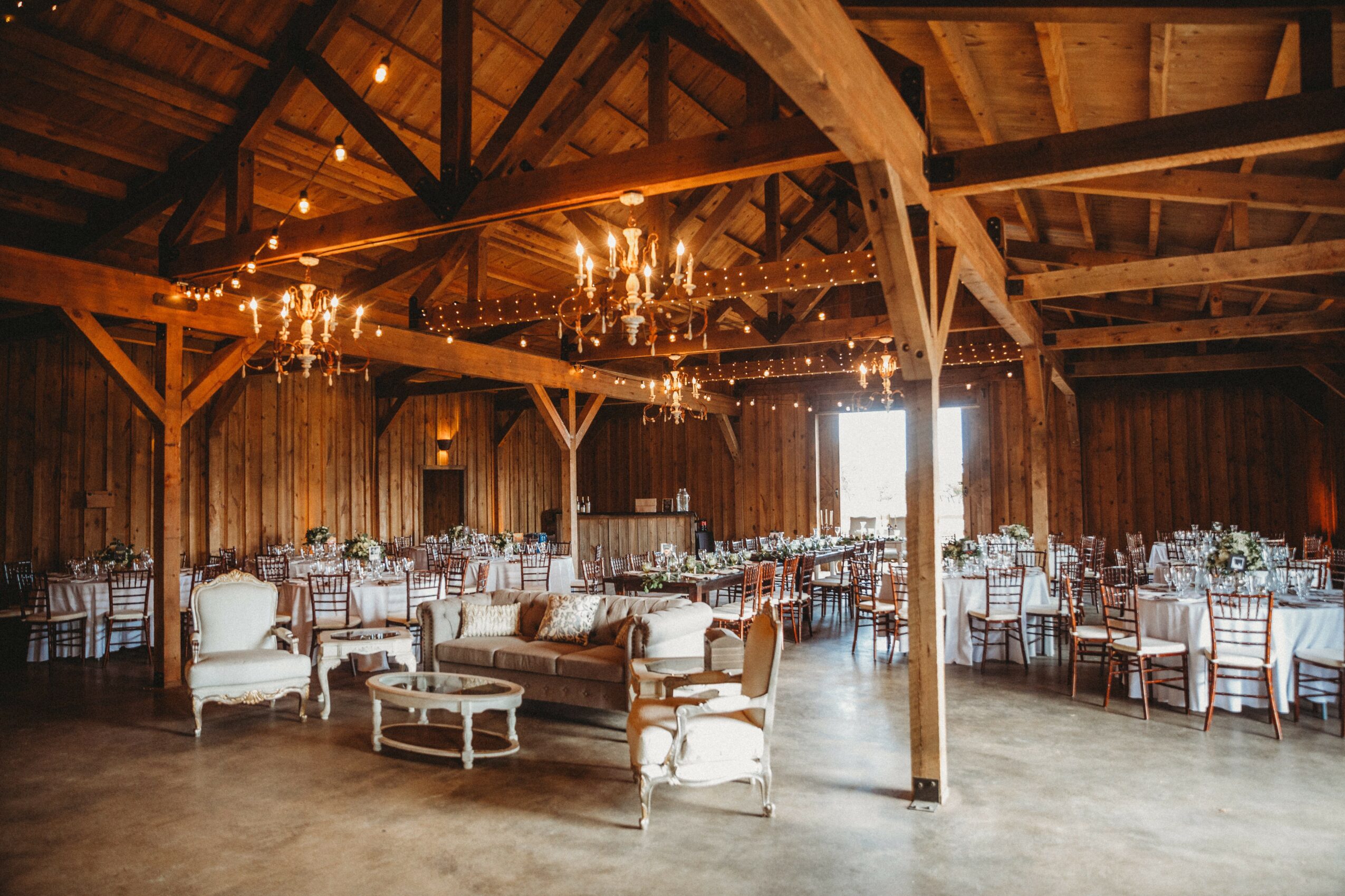 Event space with wooden walls and ceiling, chandeliers, and arranged tables and chairs, illuminated by natural and warm lighting.