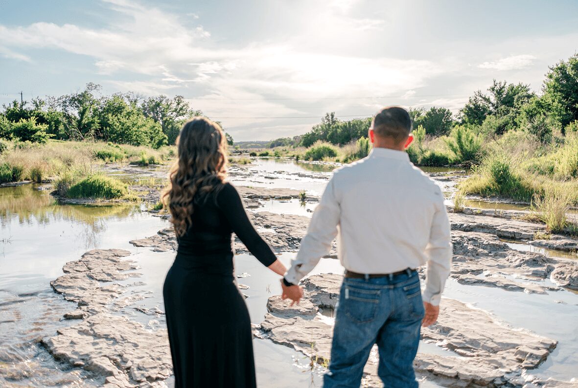 A couple holding hands by a river with rocks and greenery, facing away from the camera, under a partly cloudy sky.