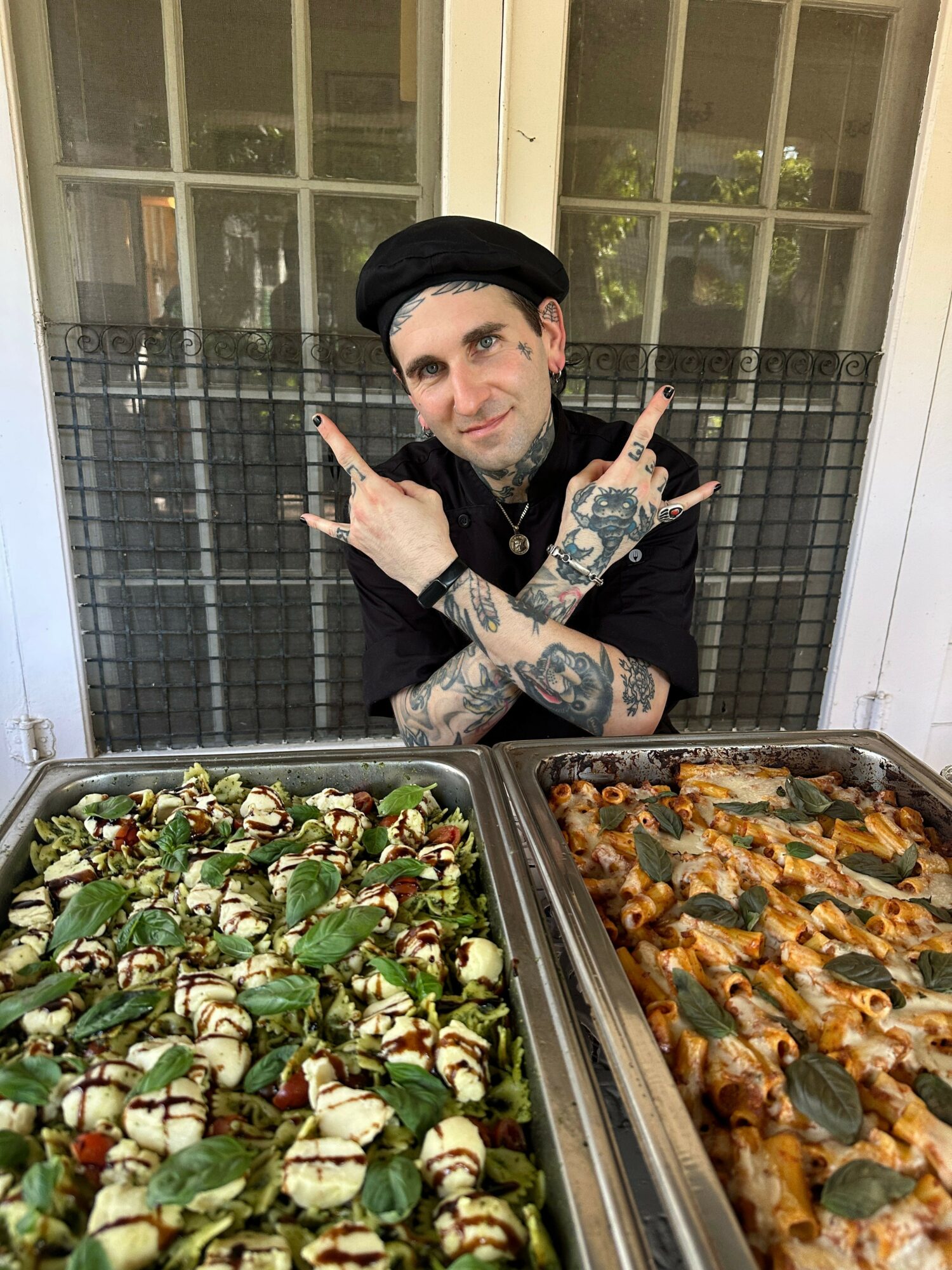 Person with tattoos, black hat, and black clothing sitting behind a metal gate, with trays of food in front.