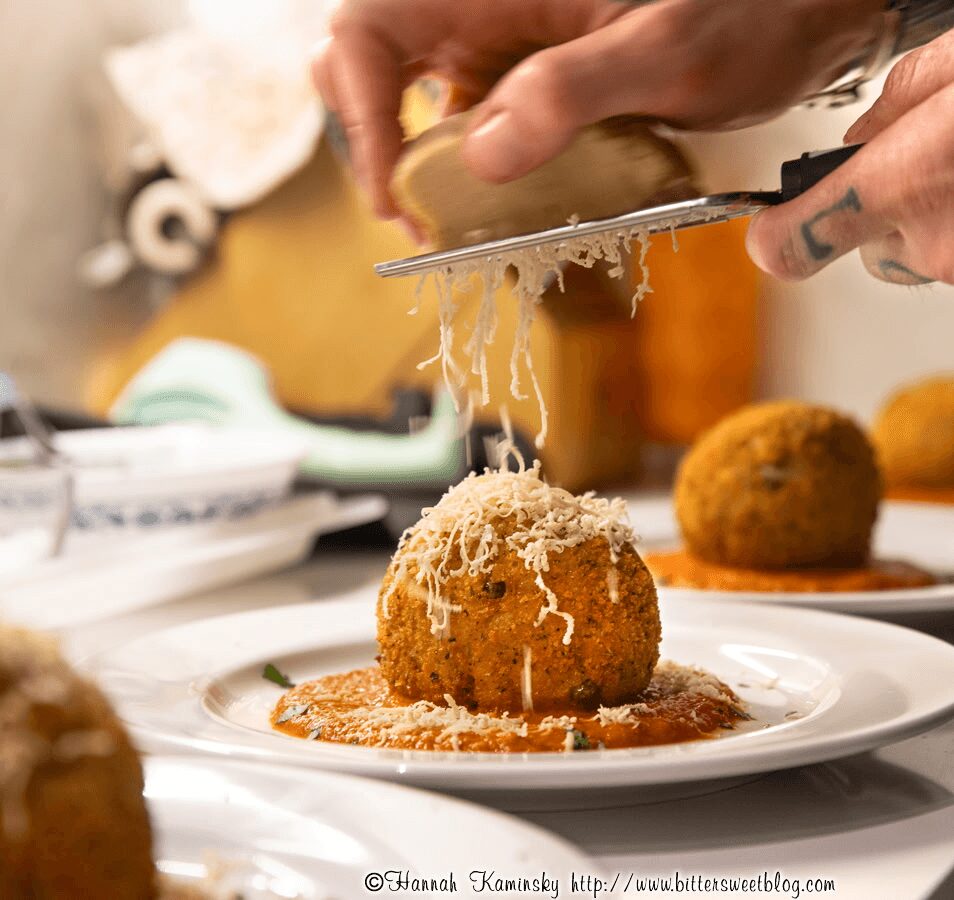 Person grating cheese over a fried ball on a white plate, with other fried balls in the background.