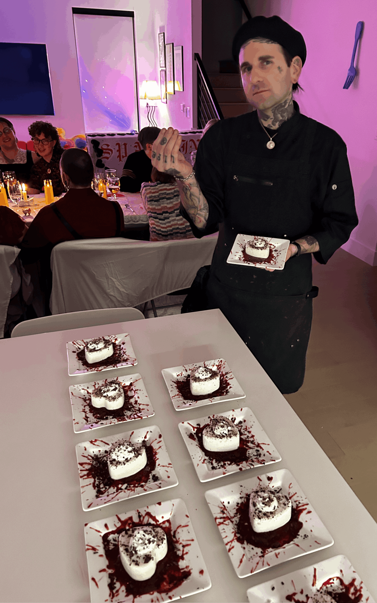 Person with tattoos and black hat holding a plate, standing behind a table with six decorated desserts, in a dimly lit room.