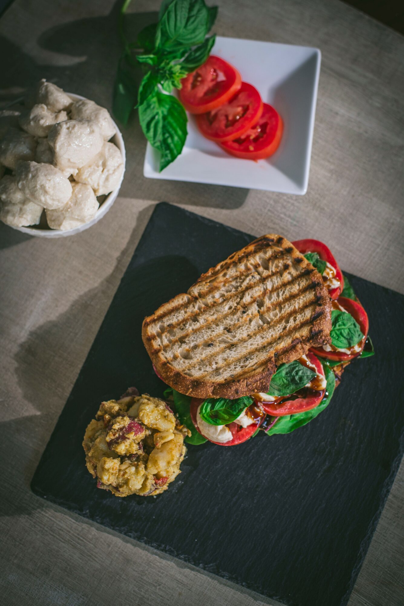 Grilled sandwich with tomatoes and basil on black slate, side of spread, bowl of cauliflower, and basil with tomato slices in white dish