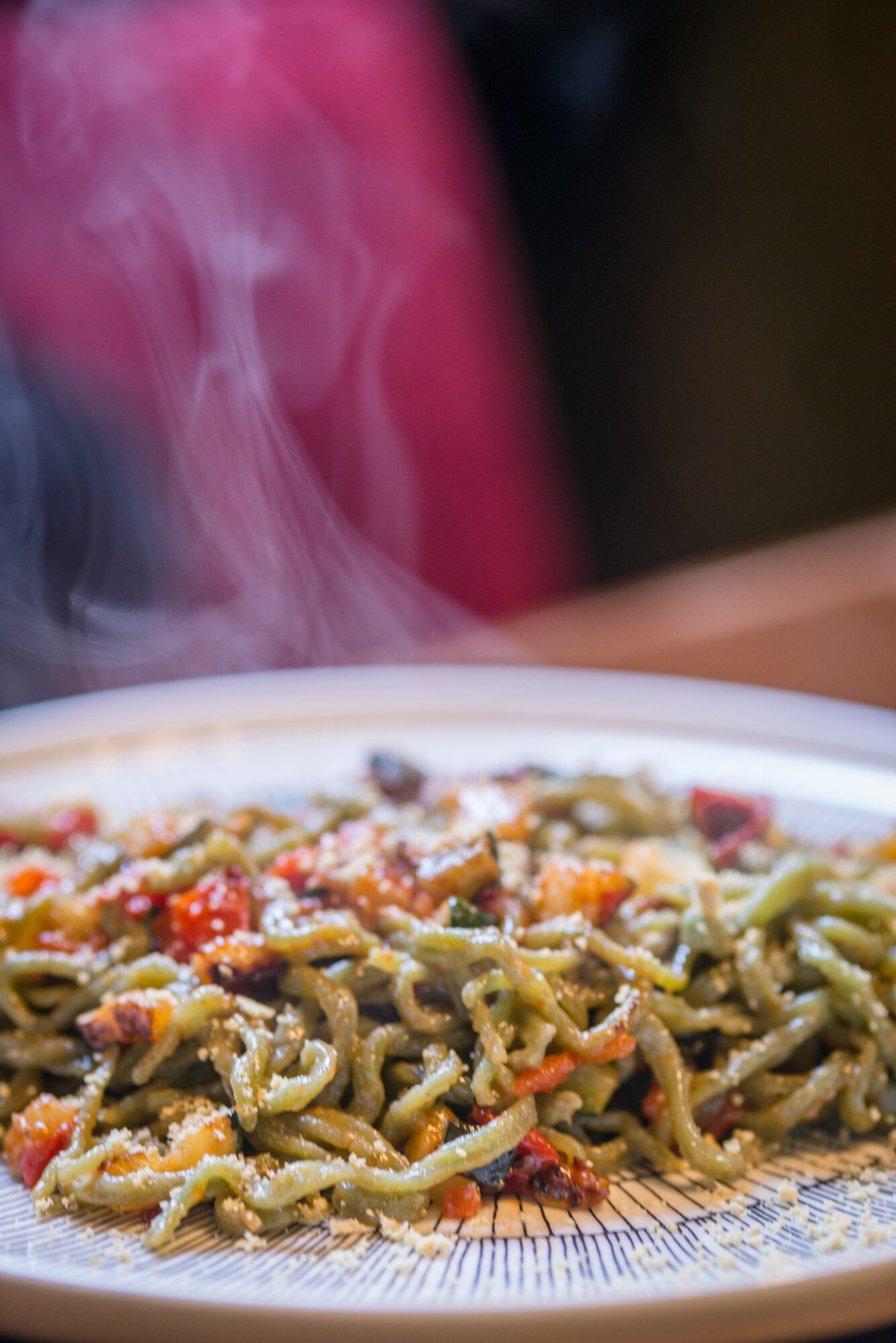 Plate of steaming pasta with vegetables on a table, with a blurred background.