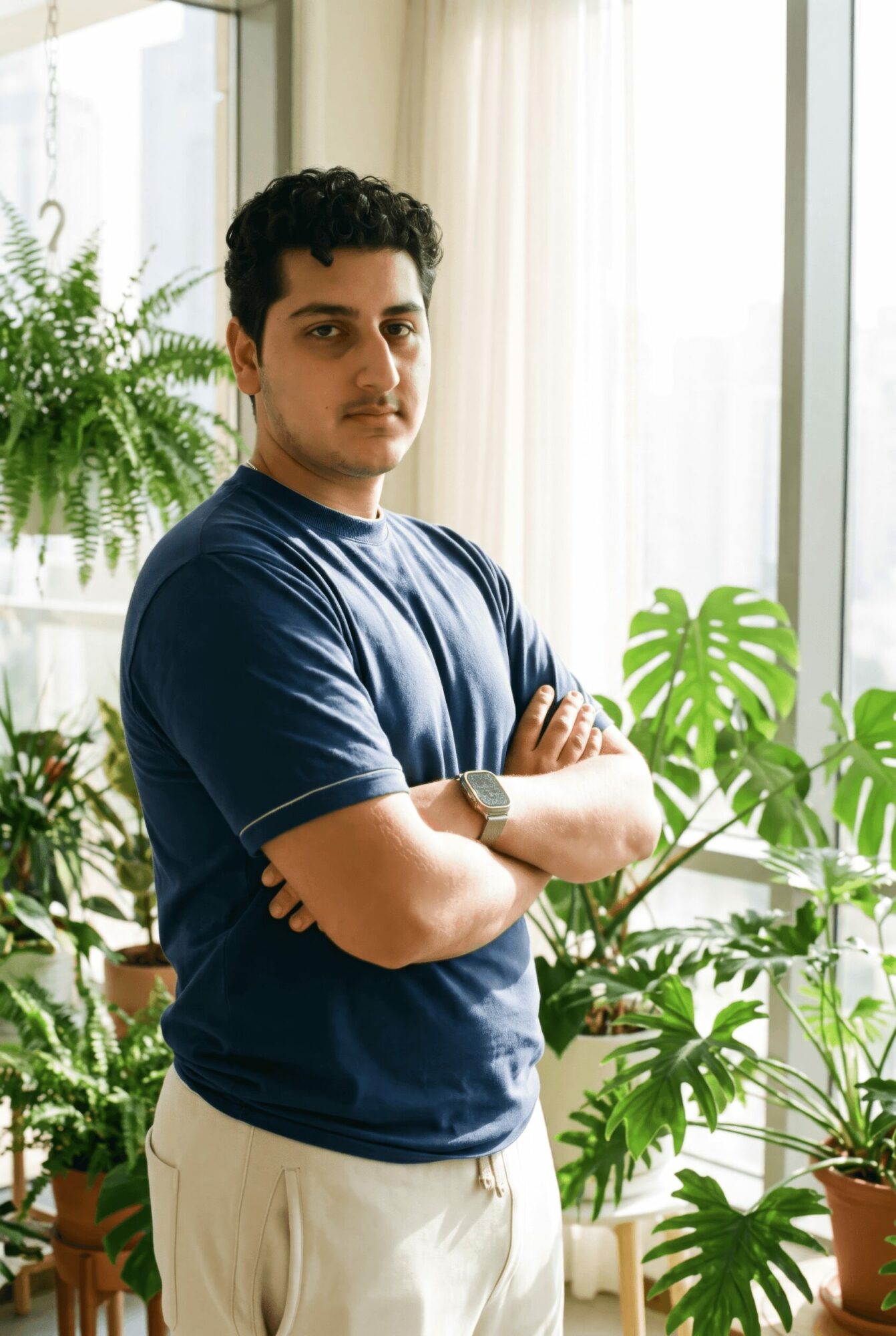 Young man with dark curly hair stands with arms crossed in a room filled with green plants, sunlight streaming through windows.