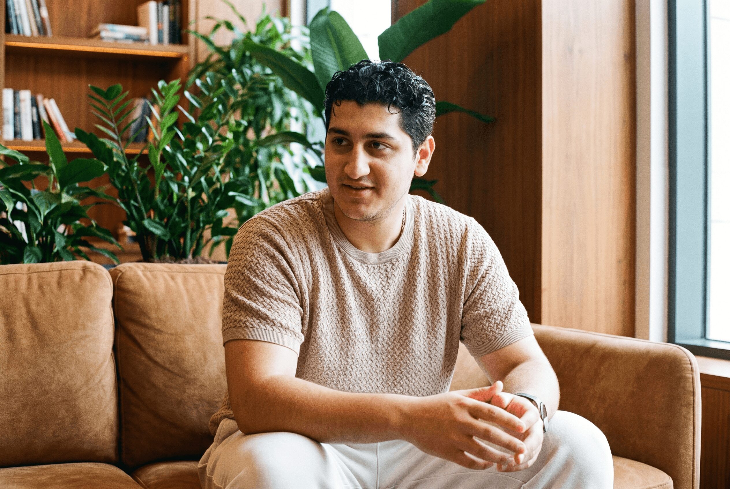 Young man sitting on a sofa in a room with plants and wooden shelves, looking to the side.