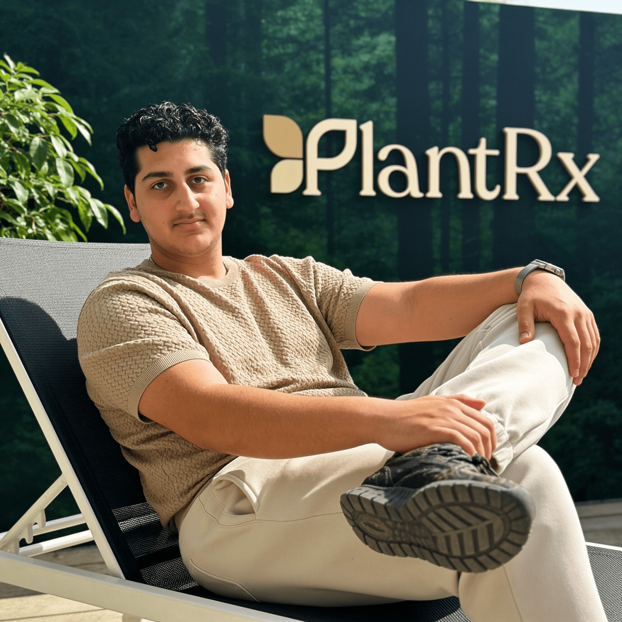 Young man sitting outdoors with a PlantRx sign in the background, wearing a beige shirt and white pants, relaxing with one leg crossed over the other.