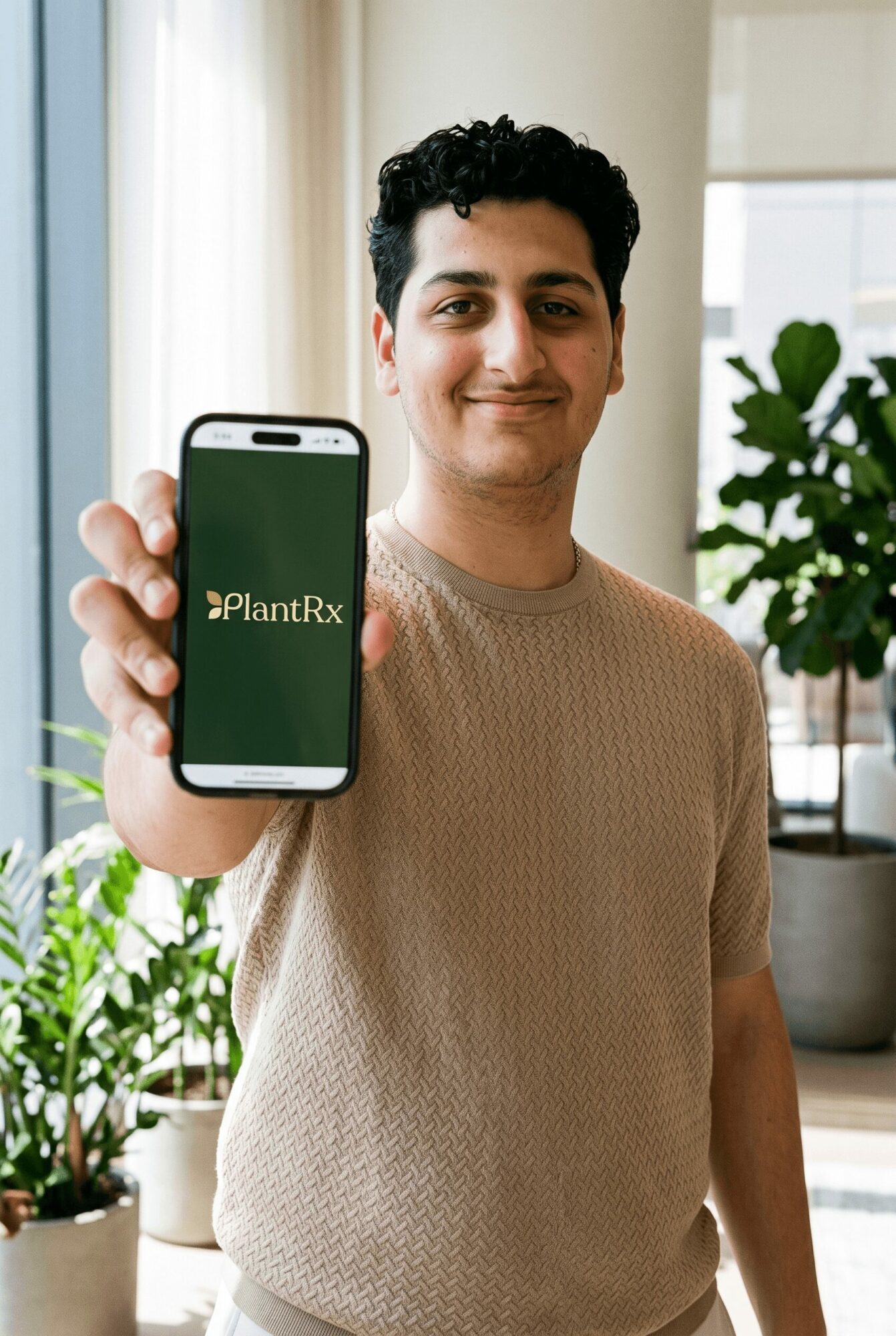 Young man holding smartphone displaying #PlantRx logo, smiling indoors with plants and large window behind him.