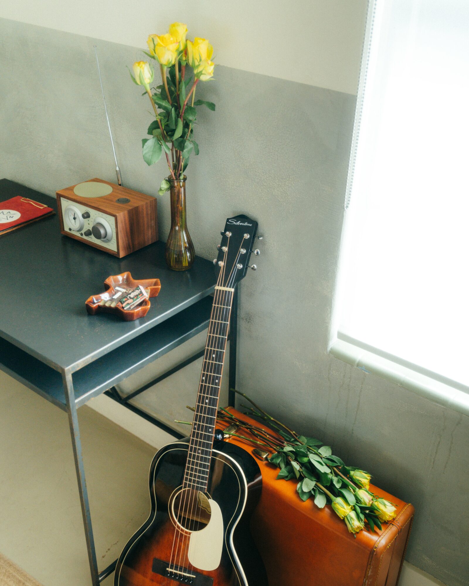 Guitar leaning against a table with a bouquet of yellow roses on top, next to a window with sunlight, and a wooden box with roses on it.