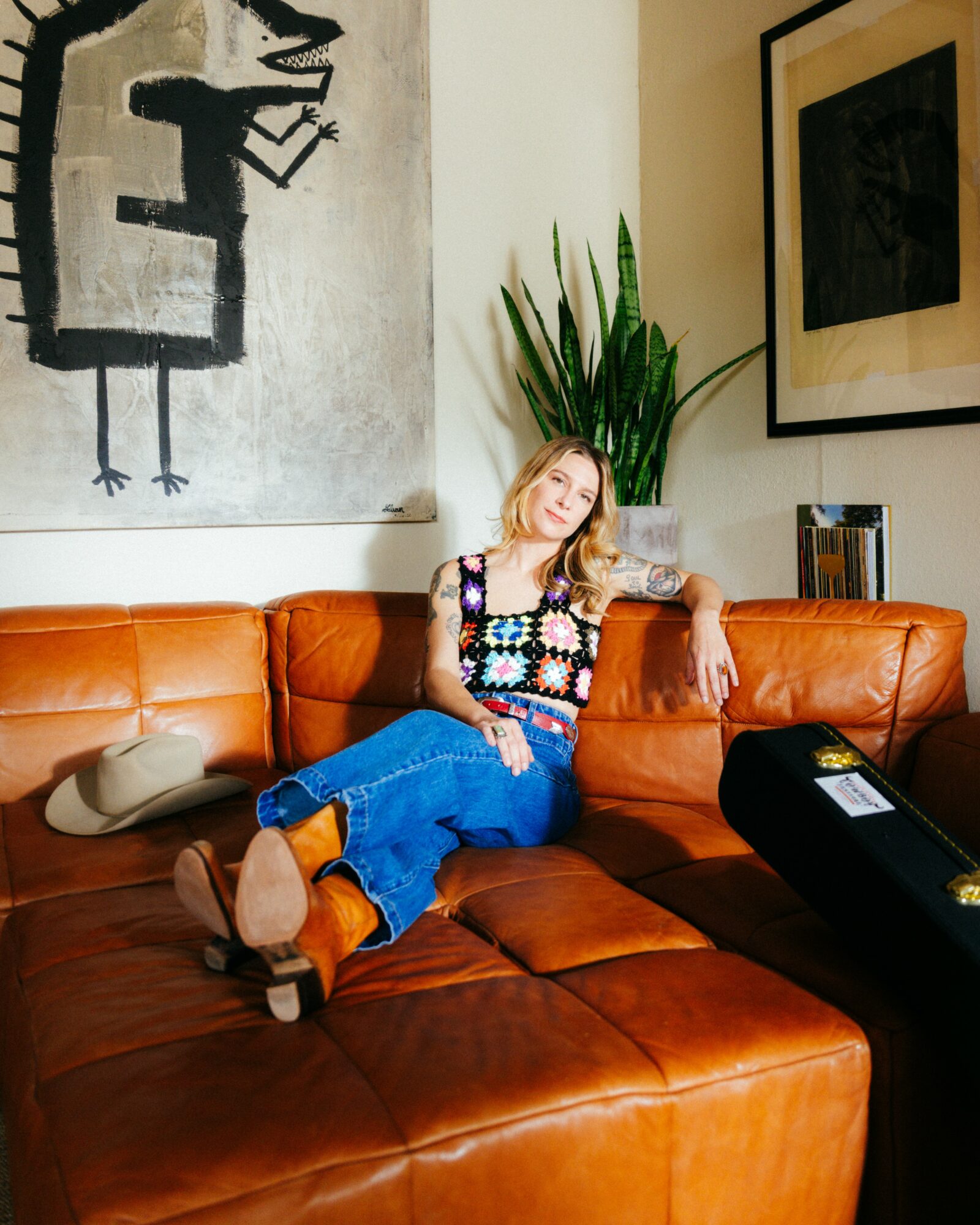 Woman sitting on a brown leather sofa, smiling, with a large plant and artwork behind her.