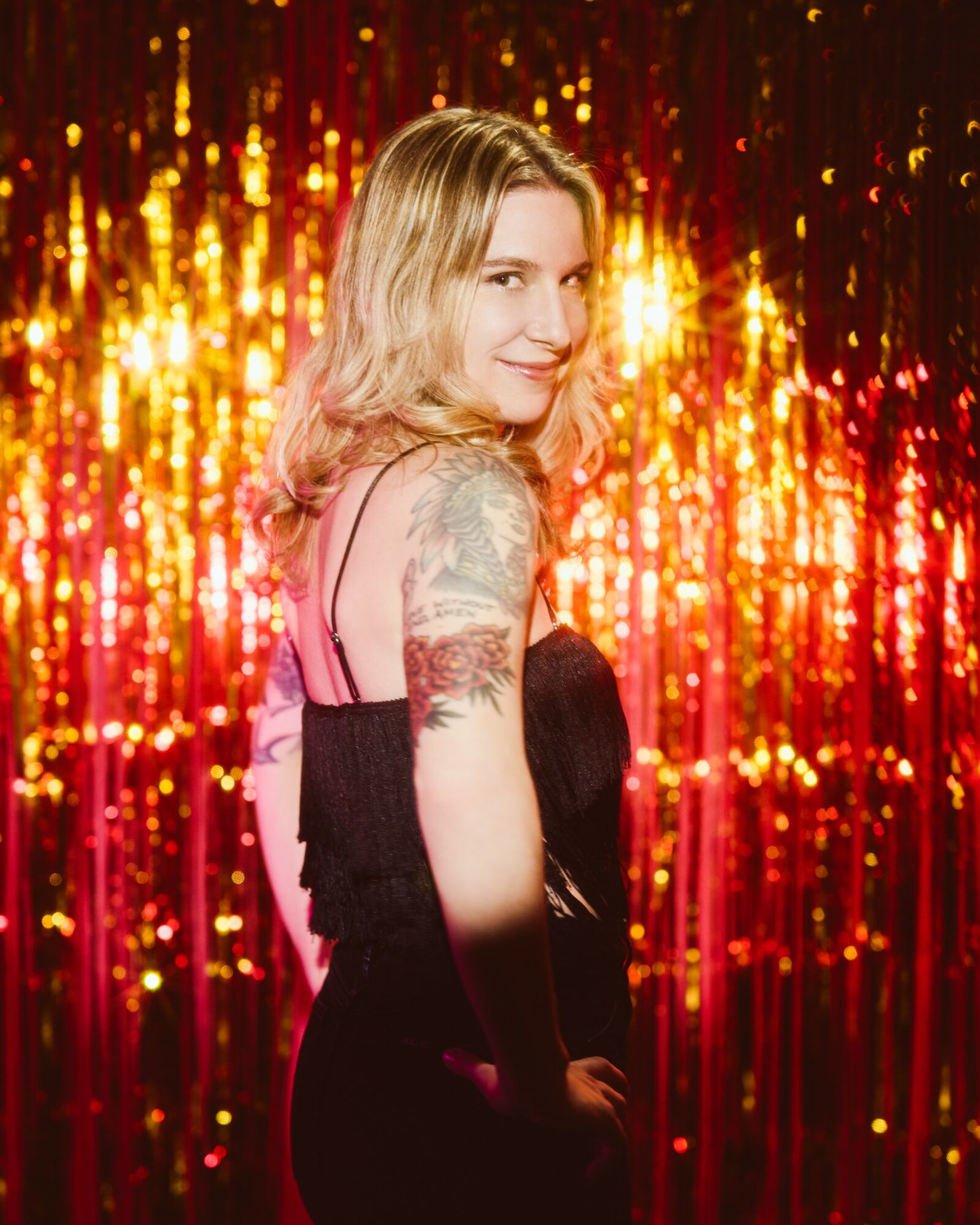 Woman with blonde hair and tattooed arm smiling in front of a gold and red glittery background.