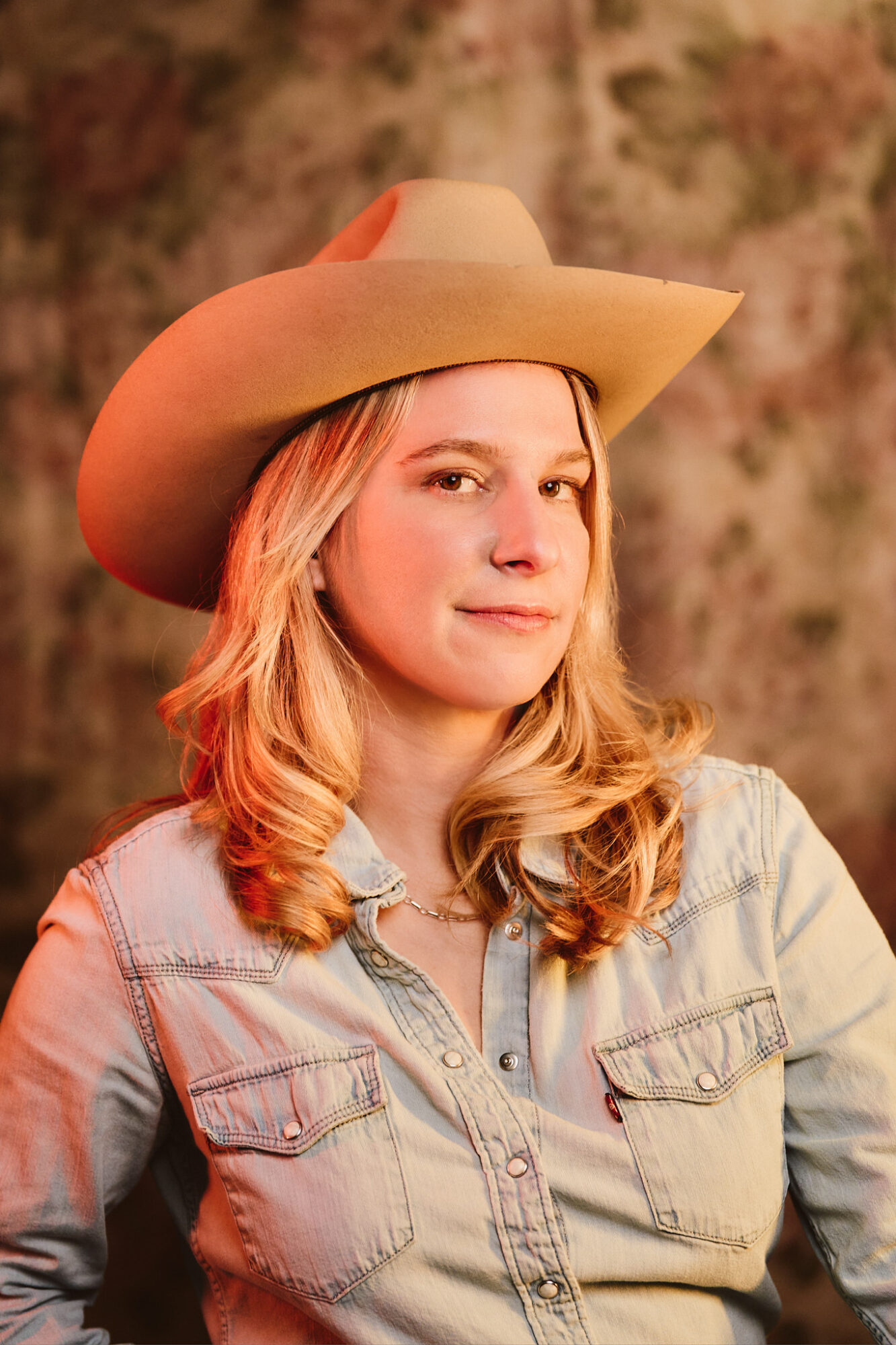 Young woman with wavy blonde hair wearing a wide-brimmed hat and denim shirt, looking at the camera.