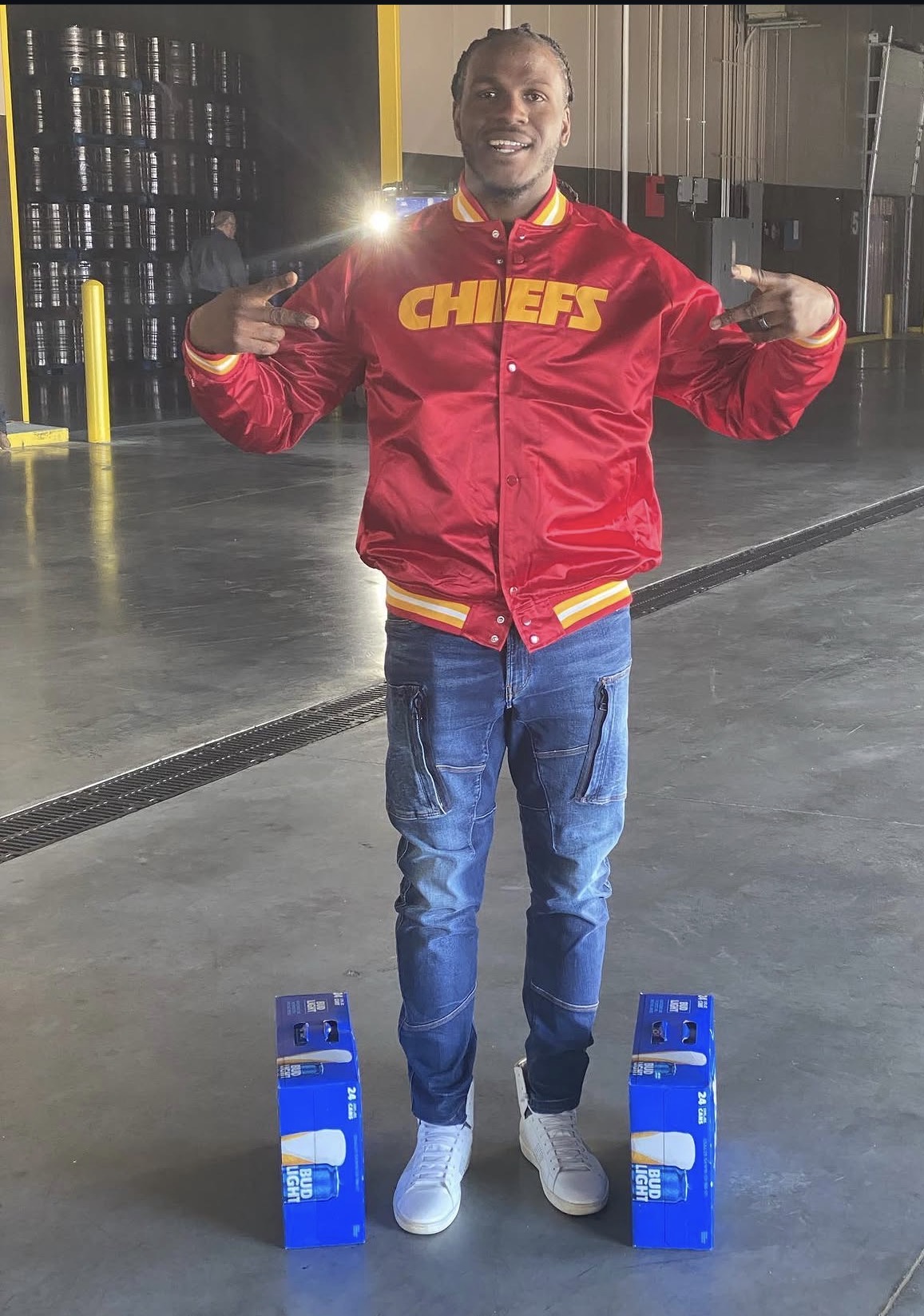 Person taking a mirror selfie in a warehouse, wearing a red Chiefs jacket, blue jeans, and white shoes, with blue objects on the floor.