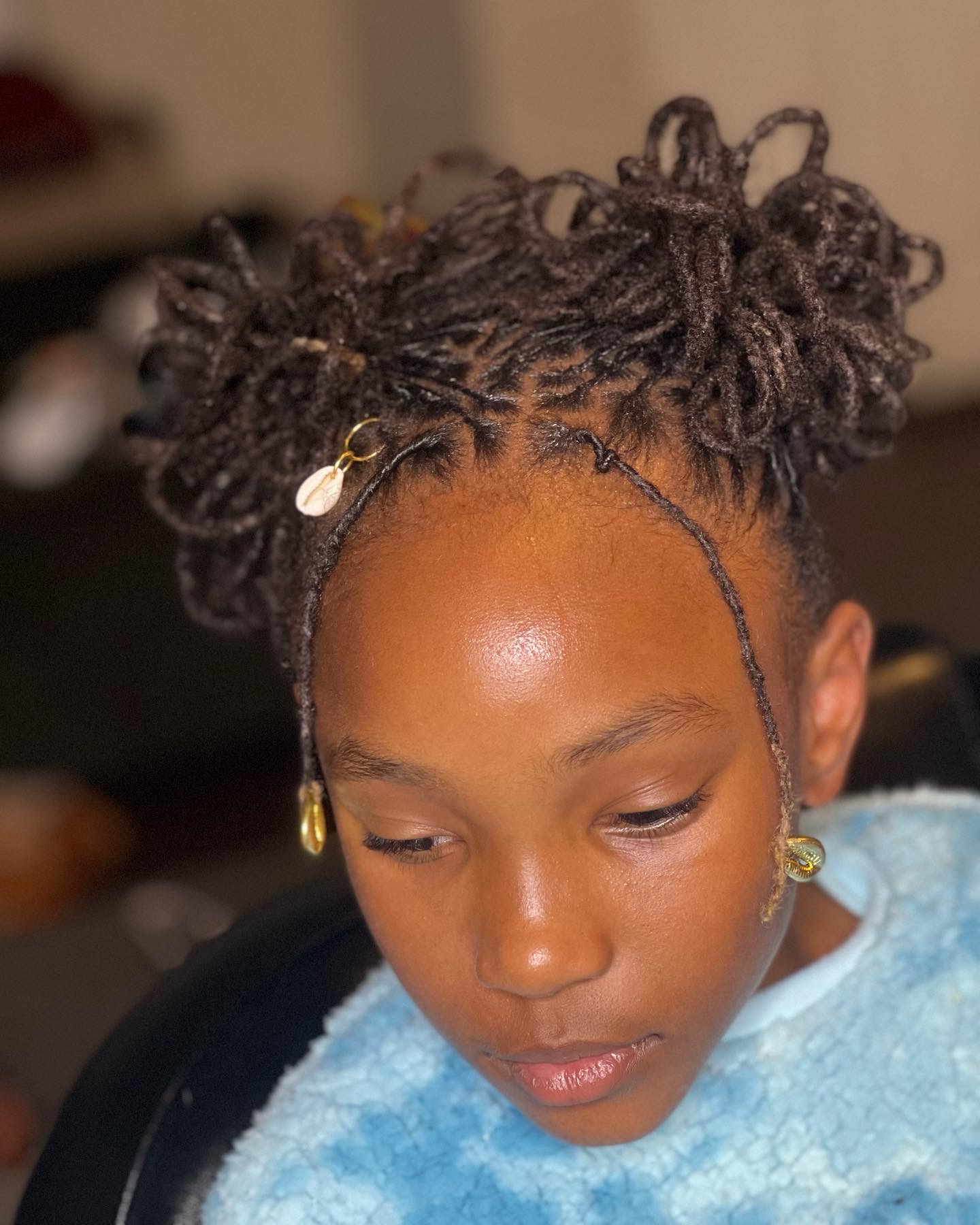 Young girl with styled dreadlocks and gold earrings, wearing a light blue garment, looking downward indoors.