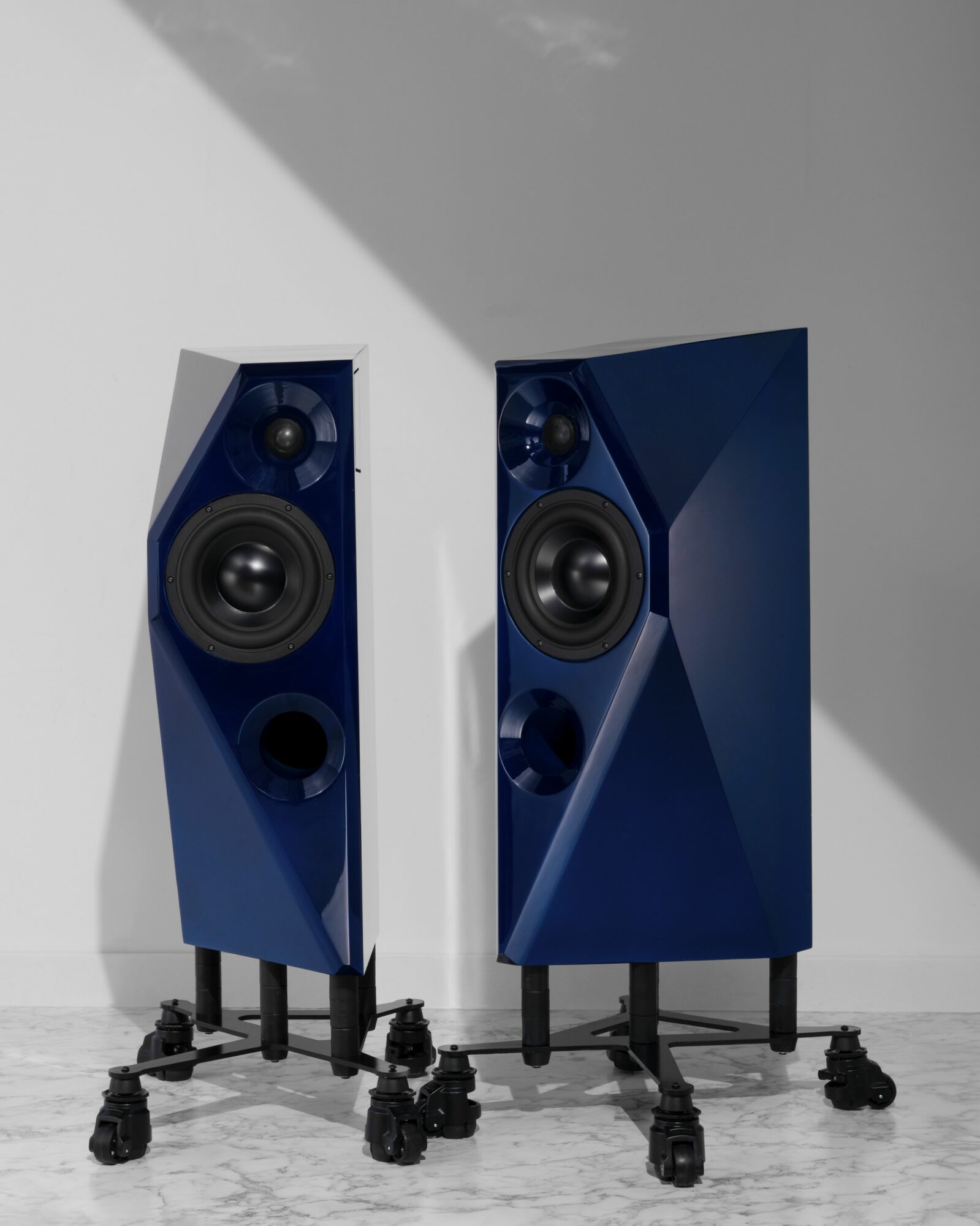 Two blue speakers on black stands against a wall, one slightly angled, with a shadow cast on the wall.