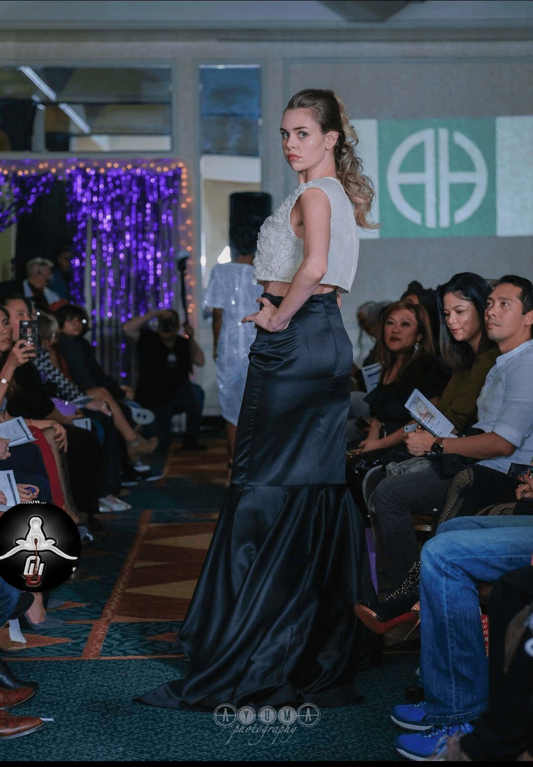 A female model walks on a runway during a fashion show, with an audience seated on both sides and decorative lighting in the background.