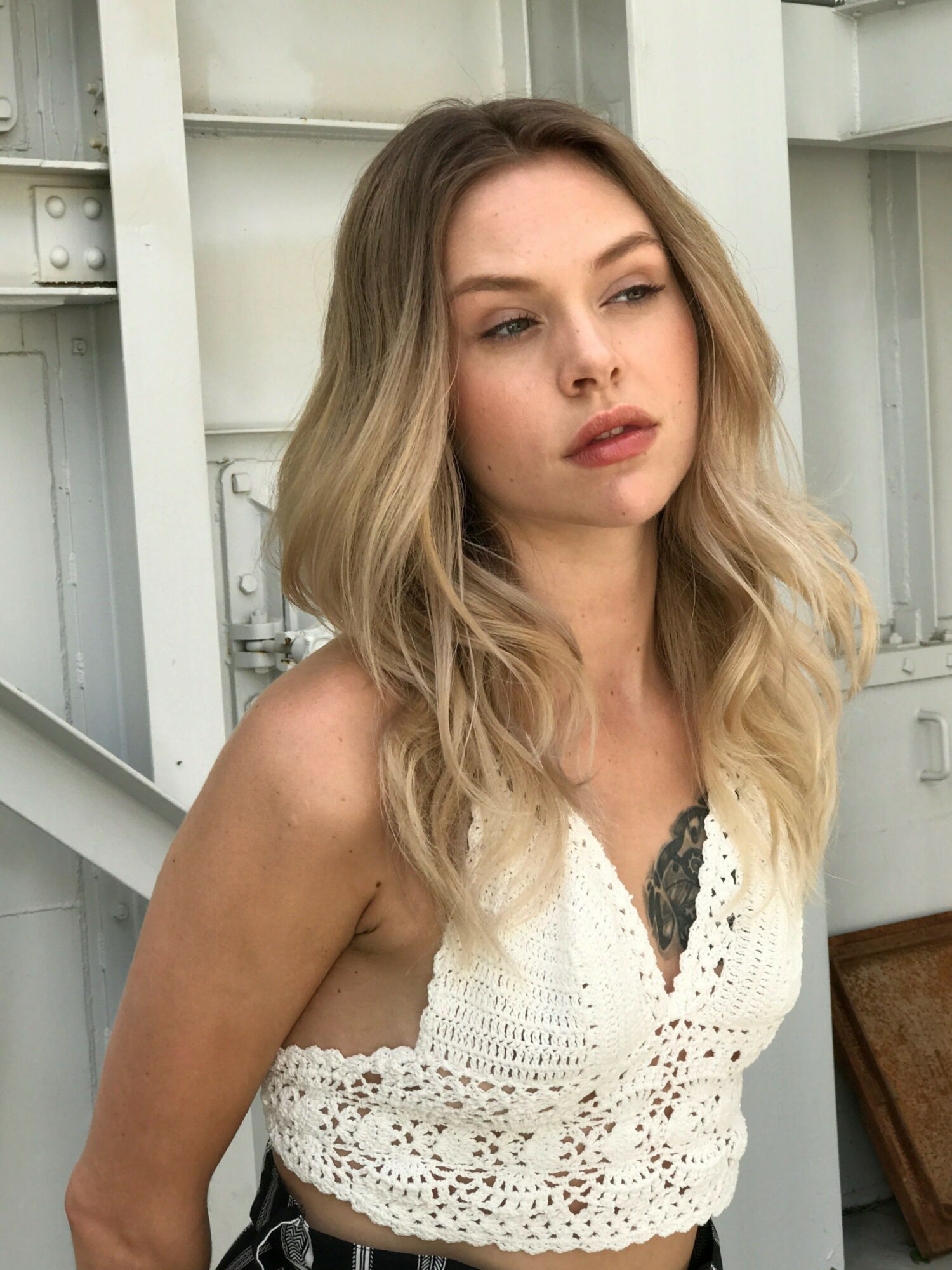 Woman with long blonde hair wearing a white crochet top, sitting indoors with a neutral expression.