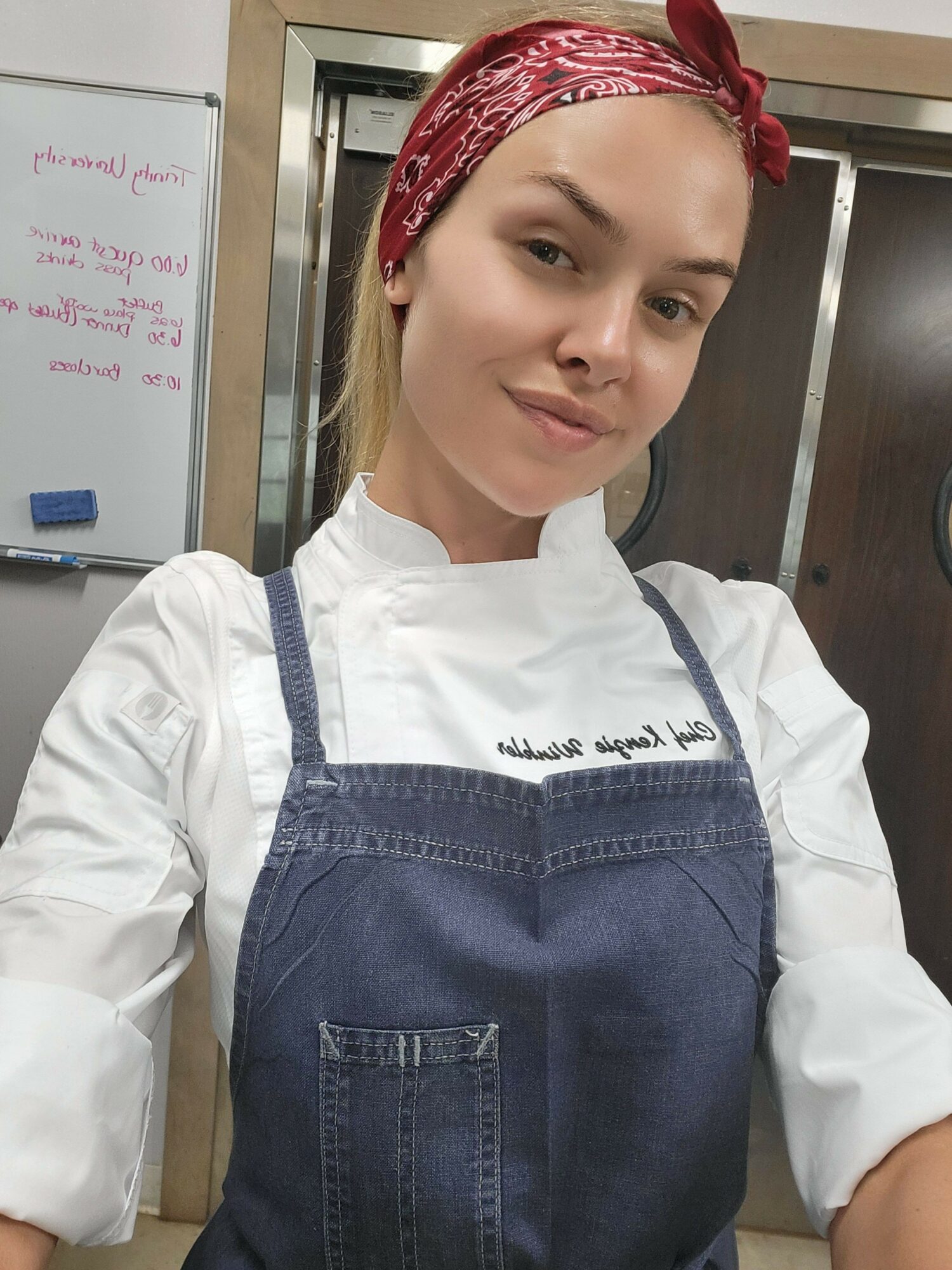 Young woman with a red headband, wearing a white shirt and denim apron, standing indoors near a whiteboard.