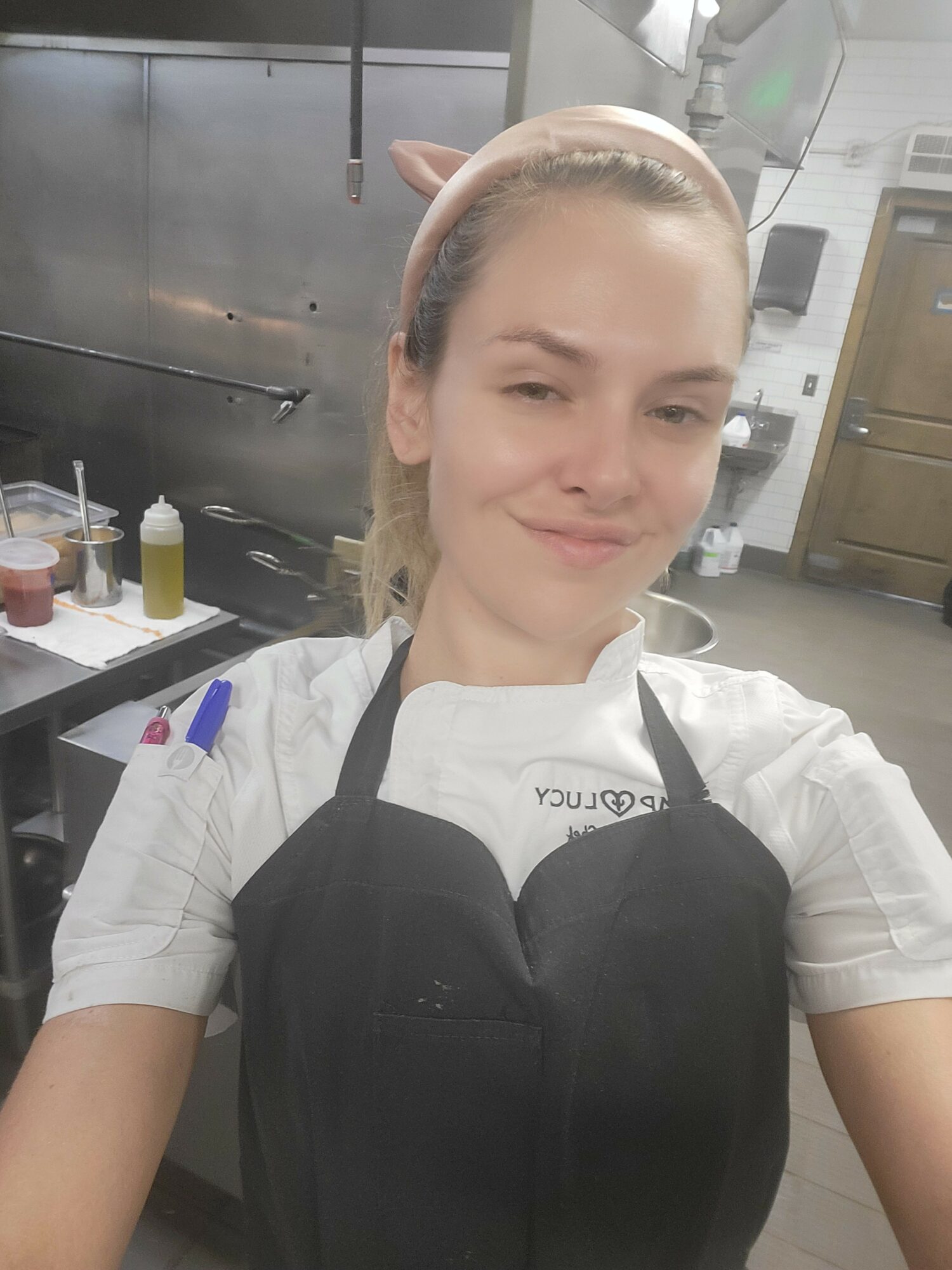 Person with elf ears and a white shirt, smiling, in a kitchen setting with appliances and utensils.