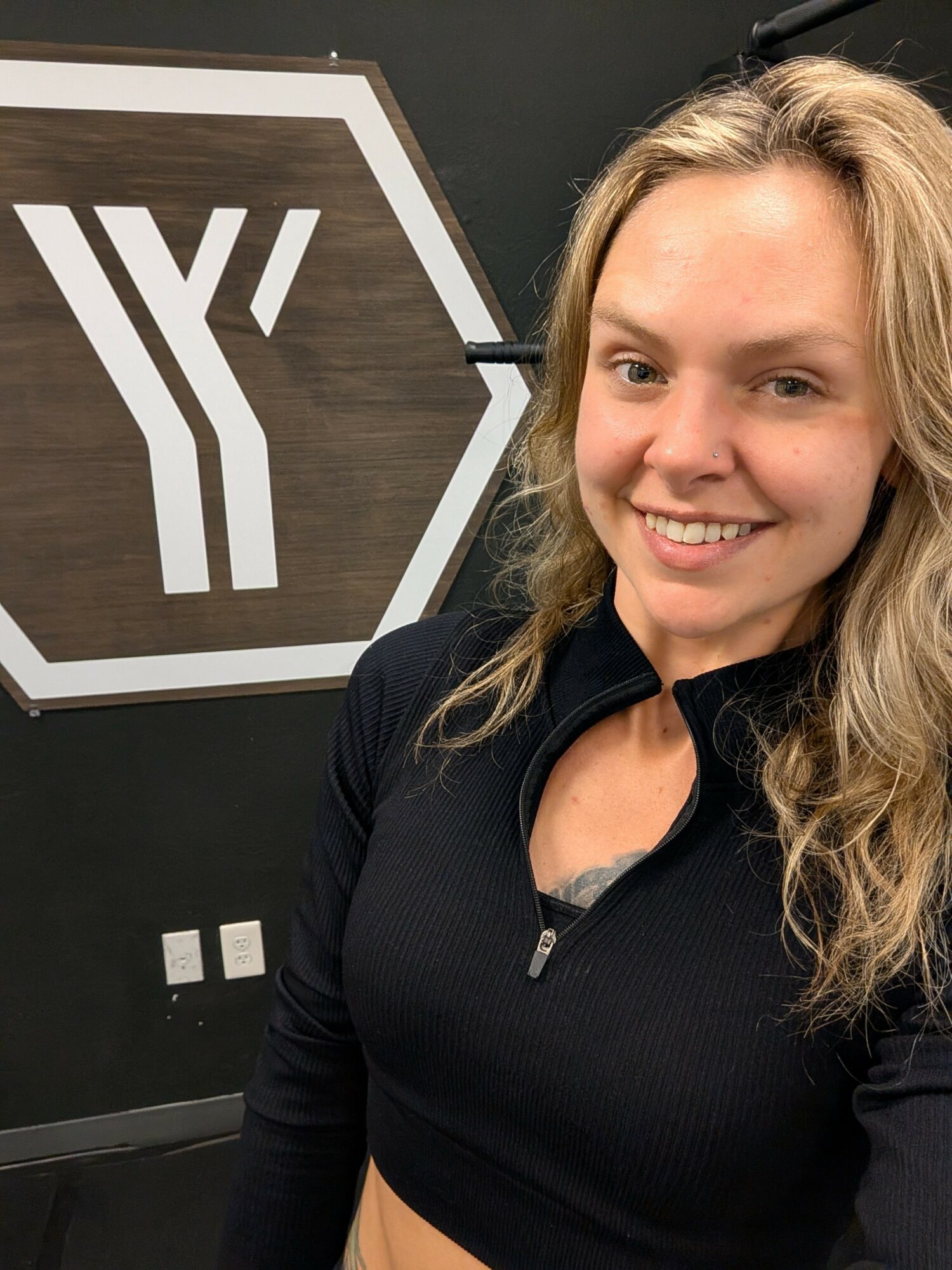 Young woman with blonde curly hair smiling, wearing a black zip-up top, standing in front of a dark background with a logo.