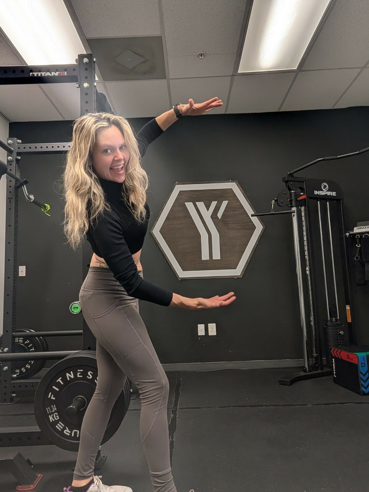 Woman with long blonde hair smiling, standing in gym, gesturing towards logo on wall, fitness equipment visible in background.