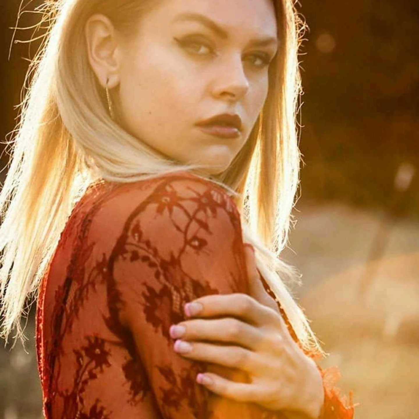 Young woman with blonde hair and dark lipstick, wearing a red lace top, looking over her shoulder outdoors during sunset.